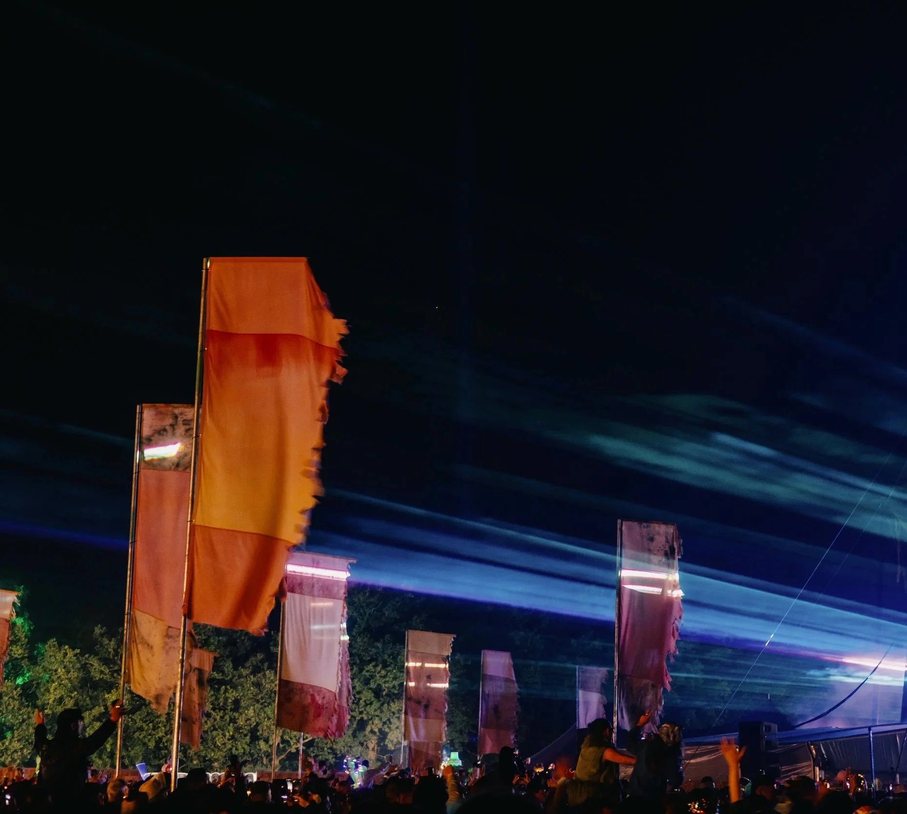 Night scene at a festival with colorful flags, bright lights, and a crowd of people.