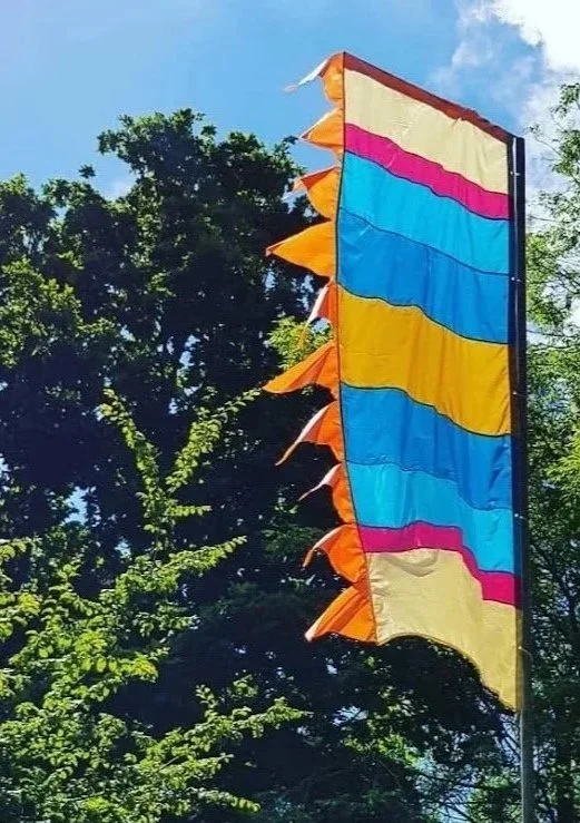 Colorful hand made bunting with pink, blue, yellow, and orange stripes flying in the breeze against a backdrop of green trees and a partly cloudy sky.