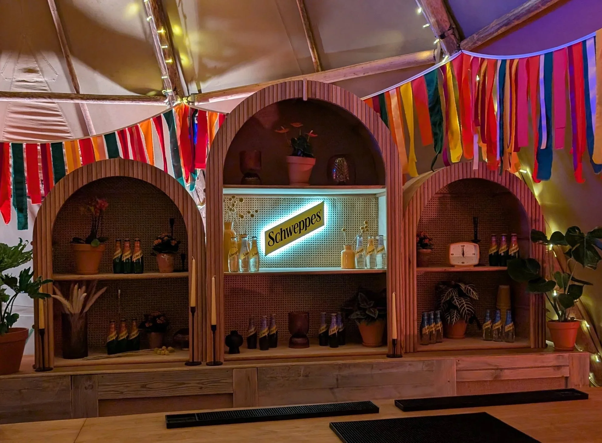Decorative bar with colorful hand made bunting, potted plants, and a Schweppes sign, illuminated with warm lighting inside a tent or canopy.