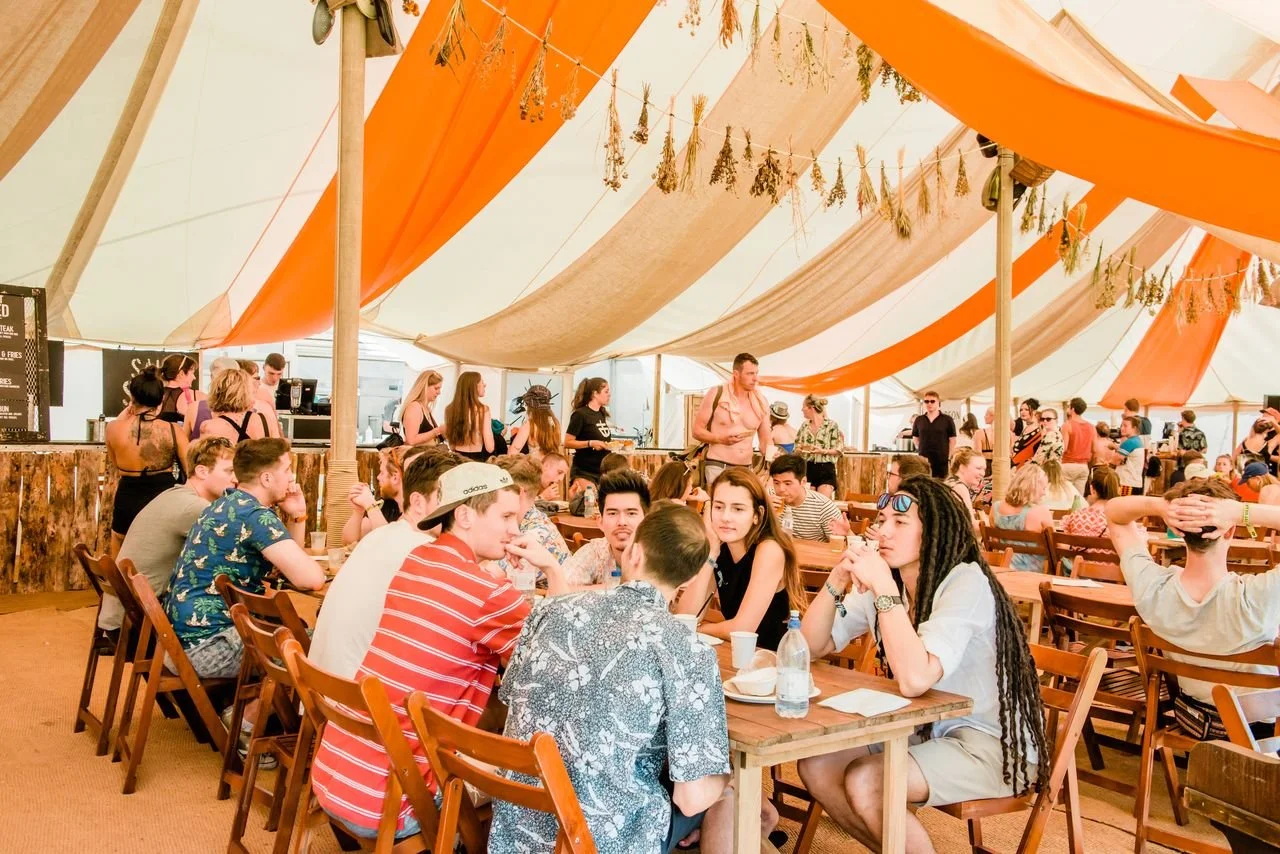 Festival attendees sitting in a dining tent at Lost Village, which is decorated with orange and hessian bunting and bunches of dried flowers