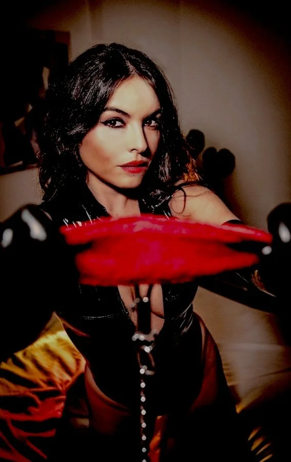 A woman with dark hair and makeup wearing black clothing holding a red object close to the camera, with a blurred background.