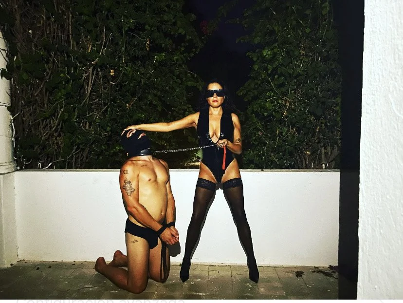 A woman in black lingerie and thigh-high stockings holding a leash on a kneeling man wearing only black briefs and a mask at night.
