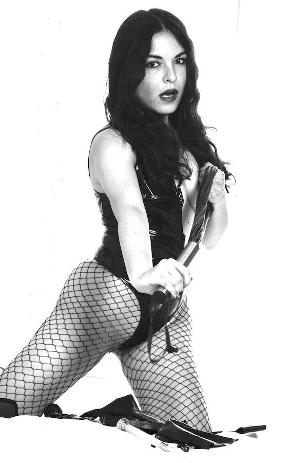 Black and white photo of a woman with long dark hair, wearing fishnet stockings and a shiny black outfit, holding a riding crop, with whips and floggers on the floor in front of her.