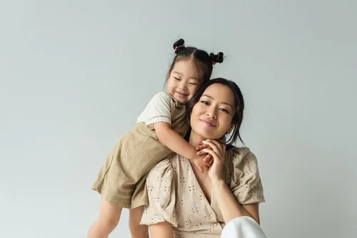 Smiling woman with a young girl hugging her from behind, both looking happy against a plain light background.