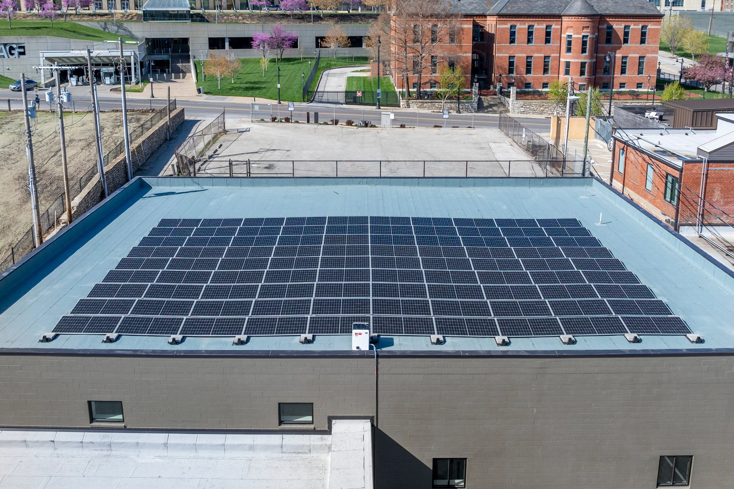 The image shows the top of a building with solar panels on the roof, with nearby buildings, trees, a street, and a parking lot in the background.
