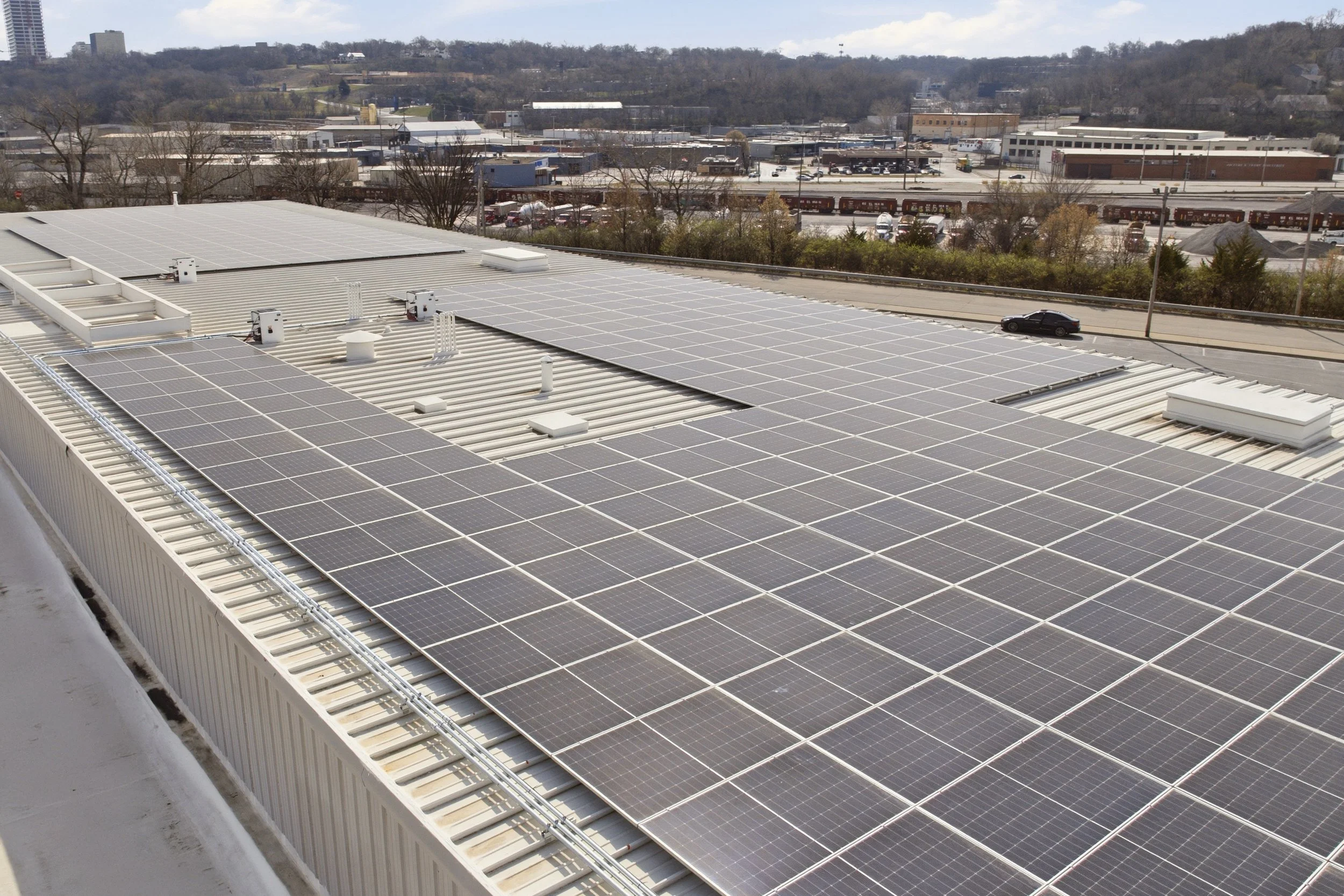 View of a rooftop in Kansas City with numerous solar panels, overlooking a parking lot and urban area with trees and buildings in the background.