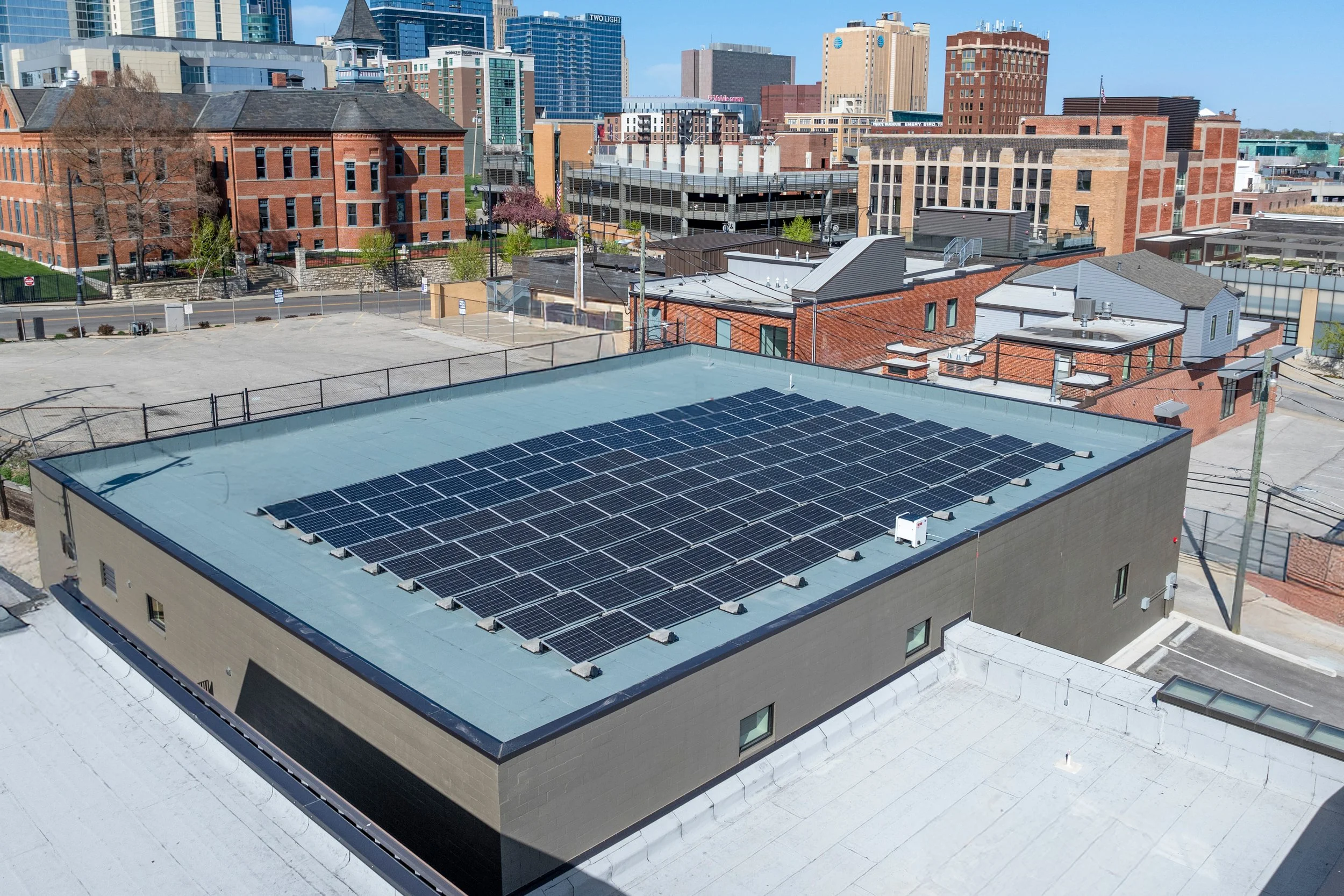 Aerial view of Kansas City with a flat building roof featuring solar panels, surrounding buildings, trees, and a clear blue sky.