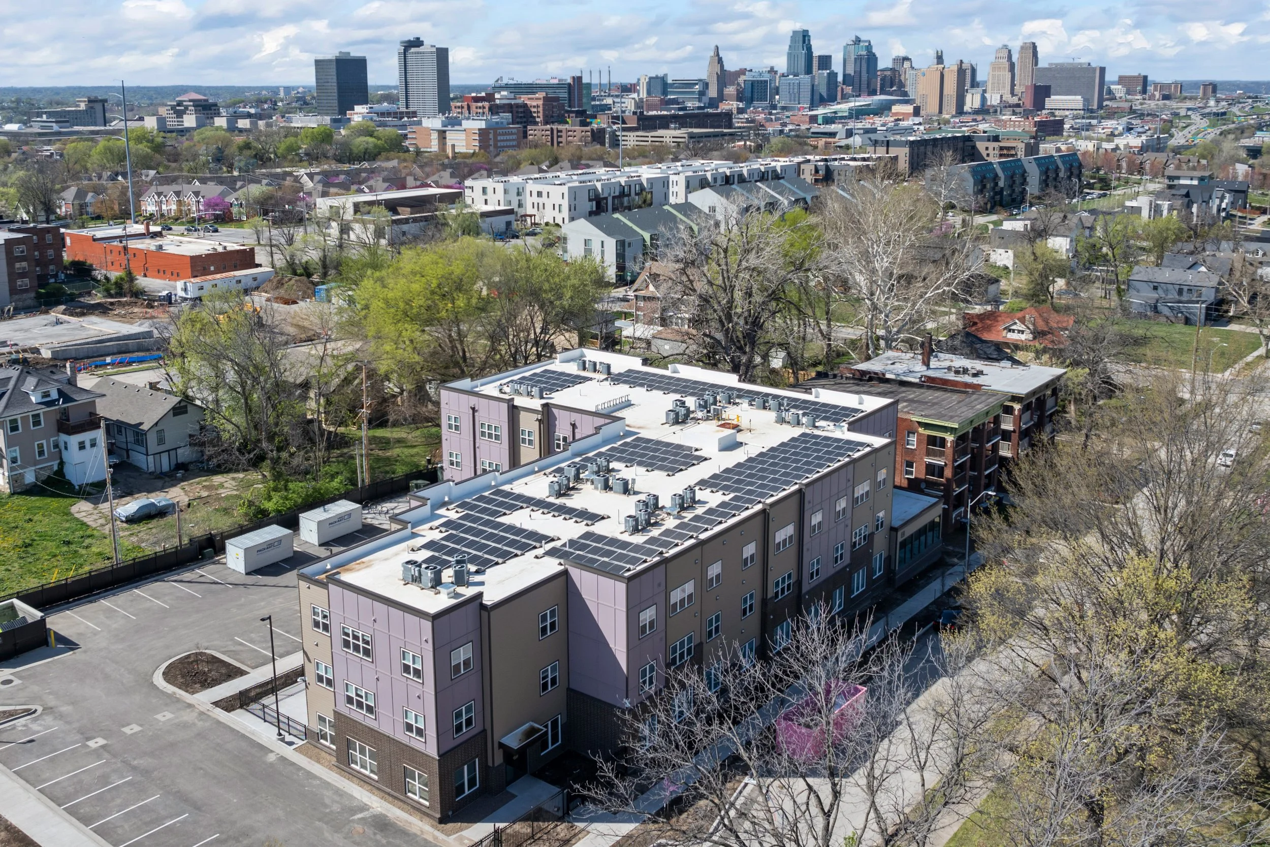 Aerial view of a Kansas City skyline with tall buildings in the background, rows of residential houses and apartment buildings in the foreground, some with solar panels on roofs, trees, and an empty parking lot.