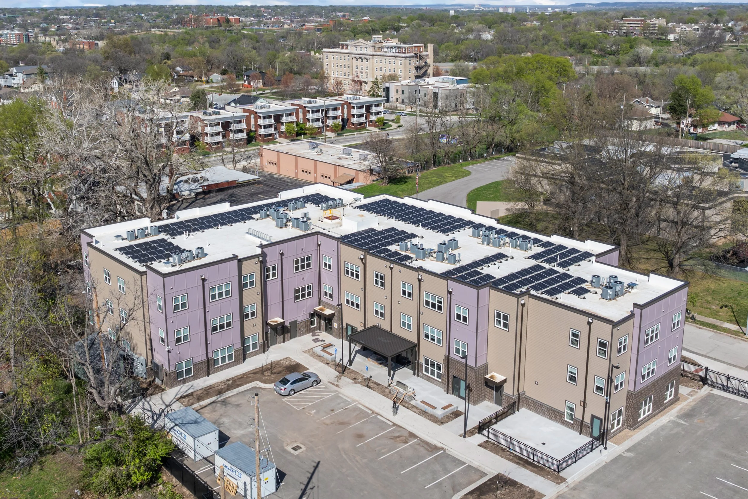Aerial view of a modern apartment complex in Kansas City, Missouri with purple-colored buildings, rooftop solar panels, and a parking lot, surrounded by trees and other residential buildings in the background.
