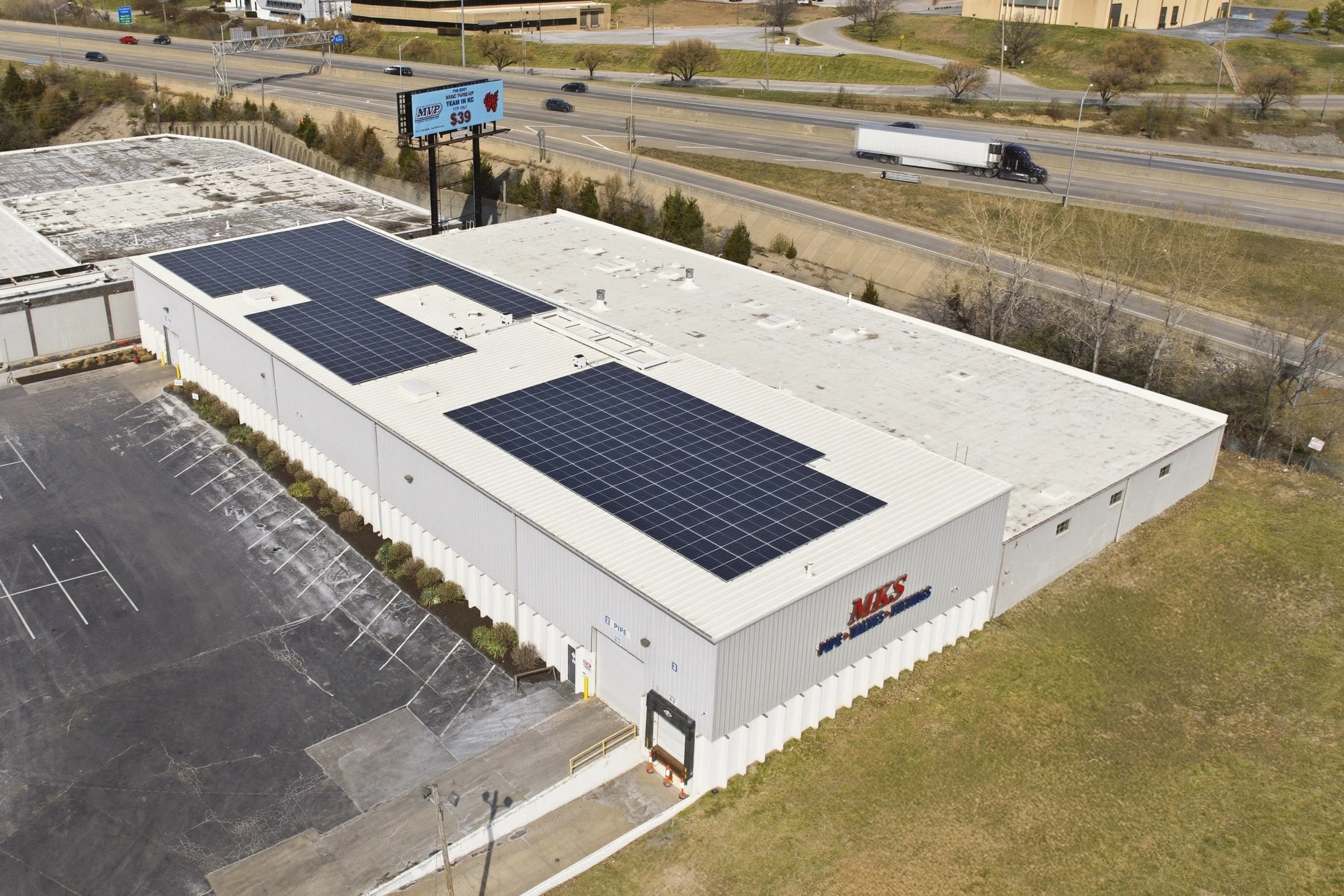 An aerial view of a large commercial building in Kansas City, Kansas with solar panels on its roof, parking lot and a busy highway with cars and trucks passing by, in the daytime.
