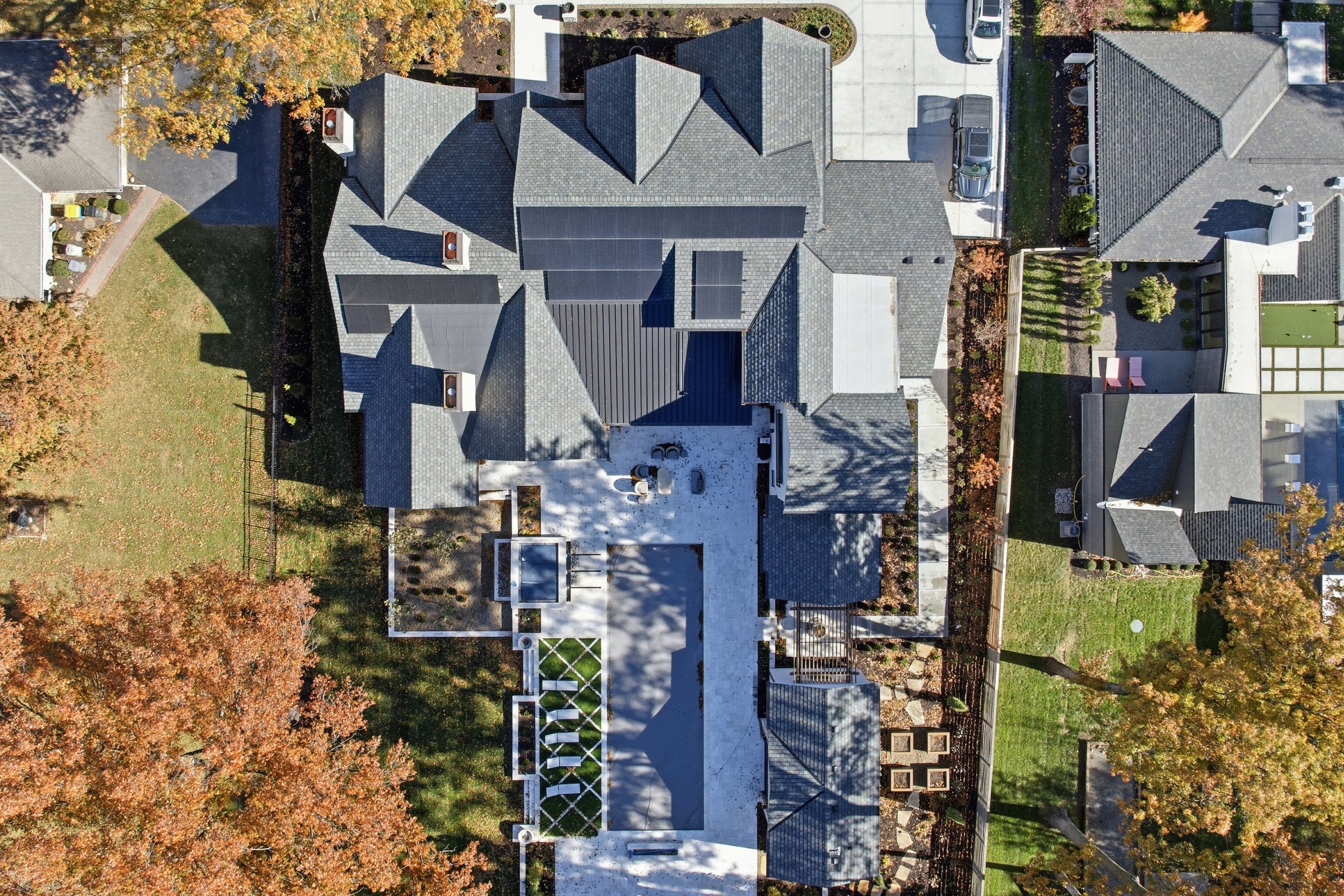 An aerial view of a residential house in Leawood, Kansas with a gray tiled roof that has 15kW of solar panels installed. The house is surrounded by trees with autumn-colored leaves, a lawn, and a paved driveway with parked cars. Sustainability. Eco
