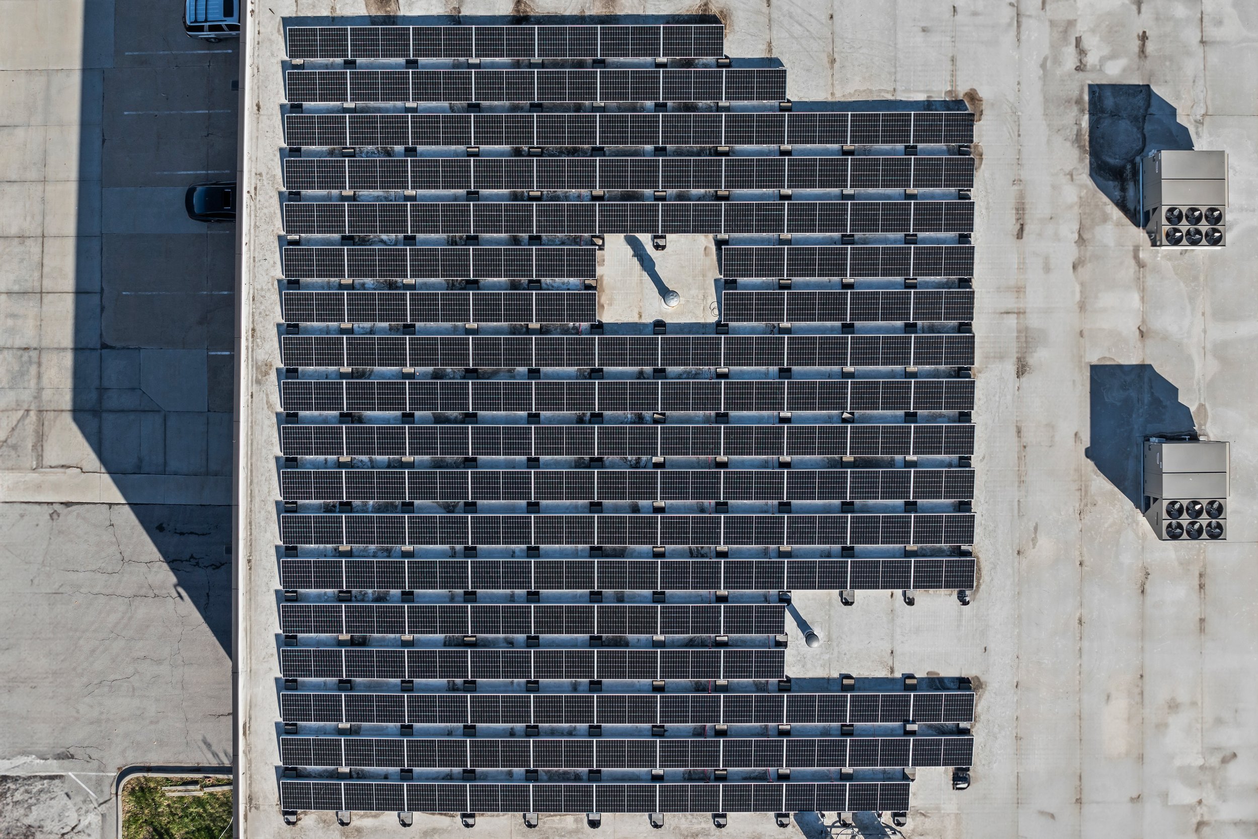 Aerial view of a rooftop with multiple rows of solar panels, some parking spaces, and HVAC units.
