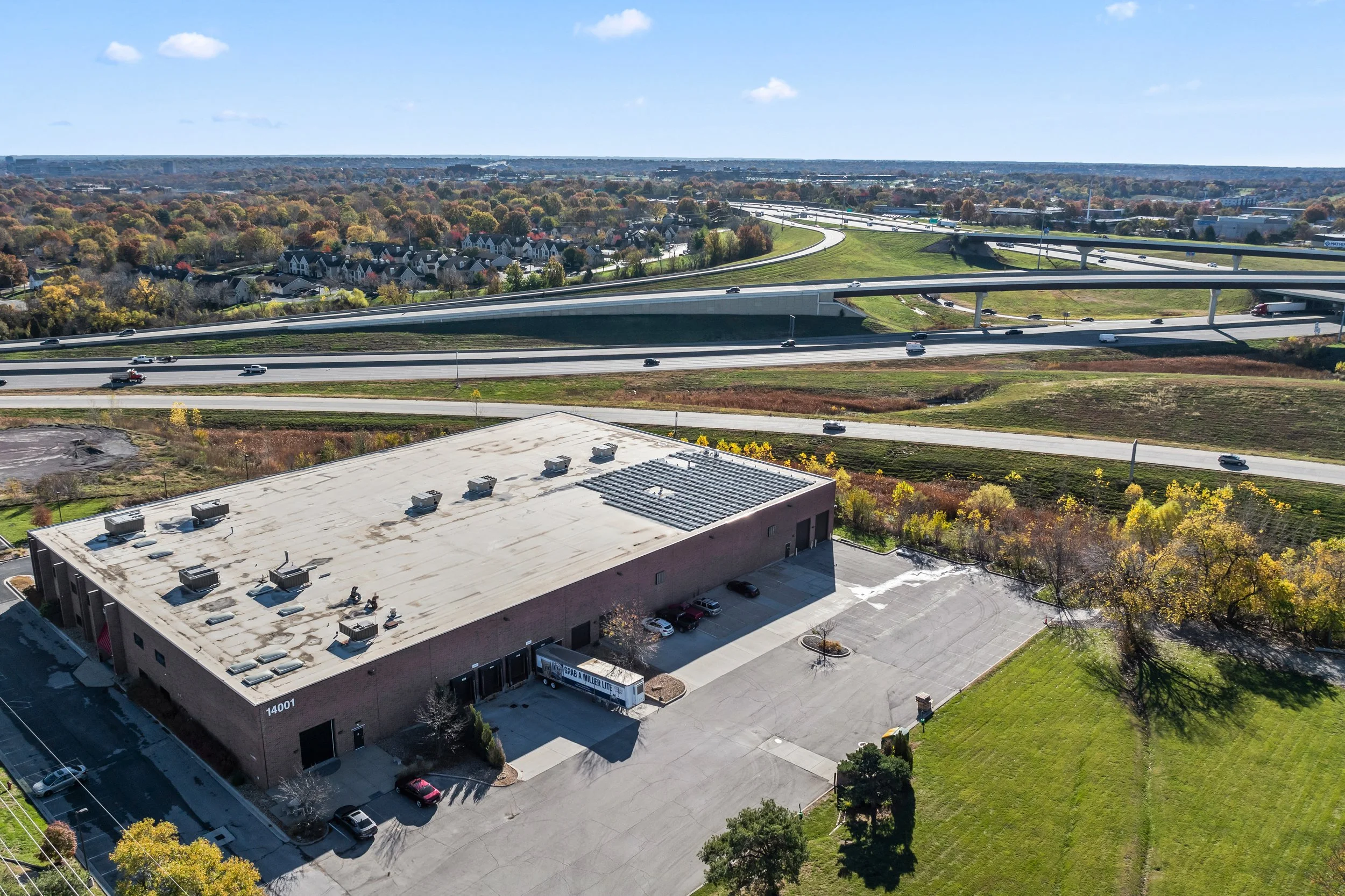 Aerial view of a cityscape showing a large commercial building in Lenexa, Kansas off of I-35 with a flat roof, multiple vehicles parked nearby, and sprawling highways with cars moving in different directions with 100kw of solar on the roof.