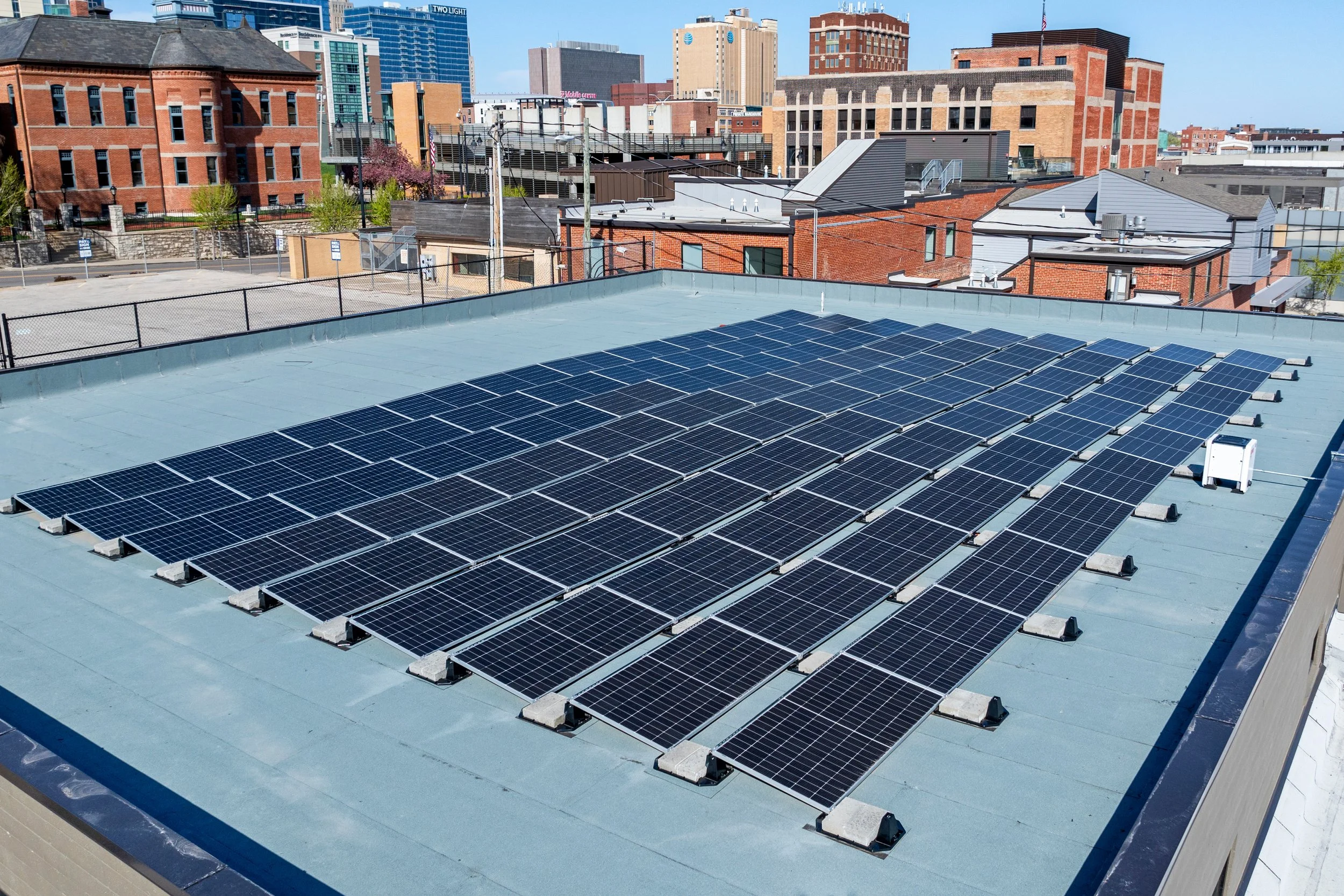 Solar panels installed on a flat rooftop of a parking garage building in an urban area with tall buildings and cityscape in the background.