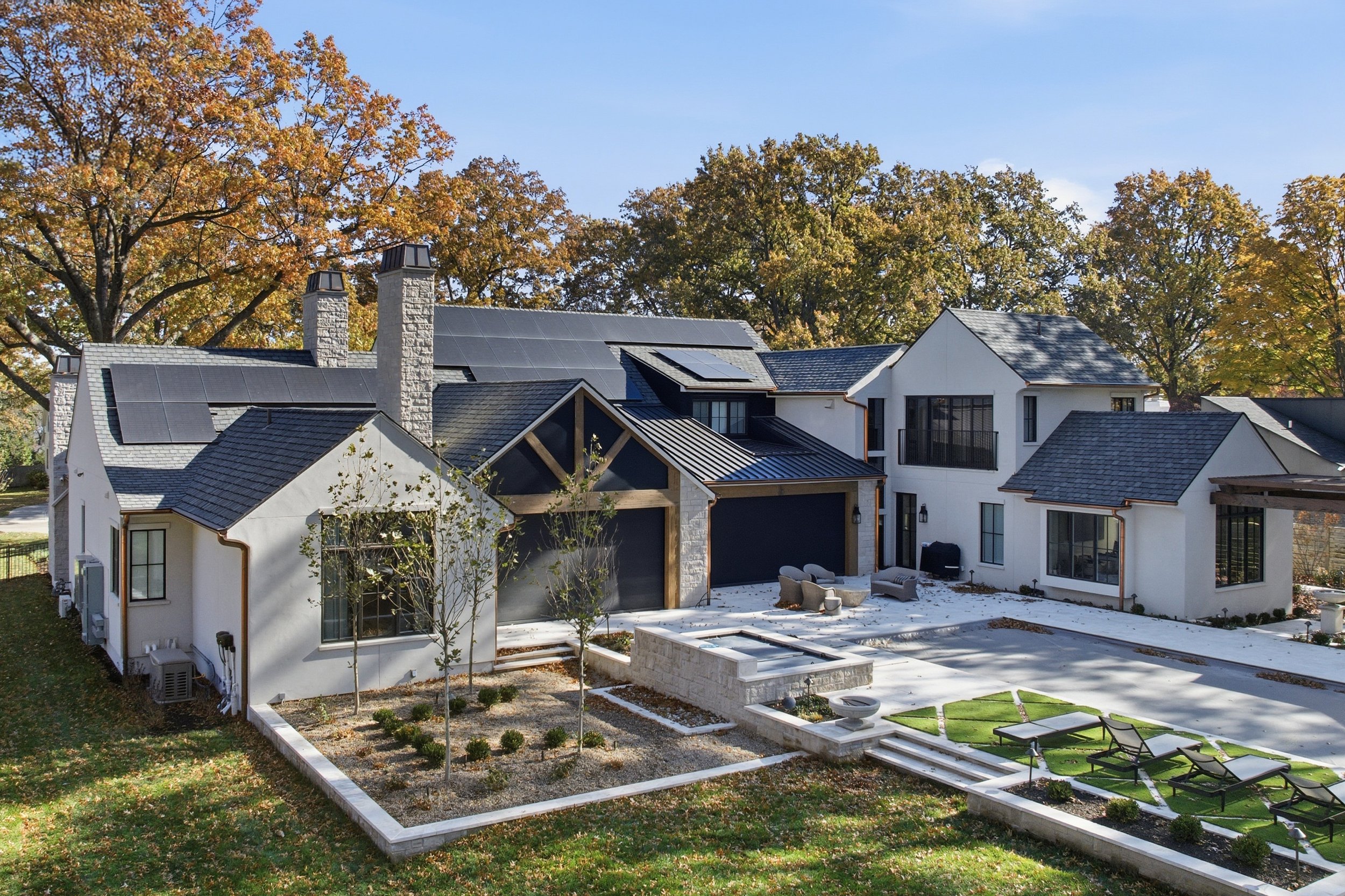 Backyard of a modern white house with solar panels on the roof, surrounded by autumn trees, a small garden with young plants, and outdoor seating area.