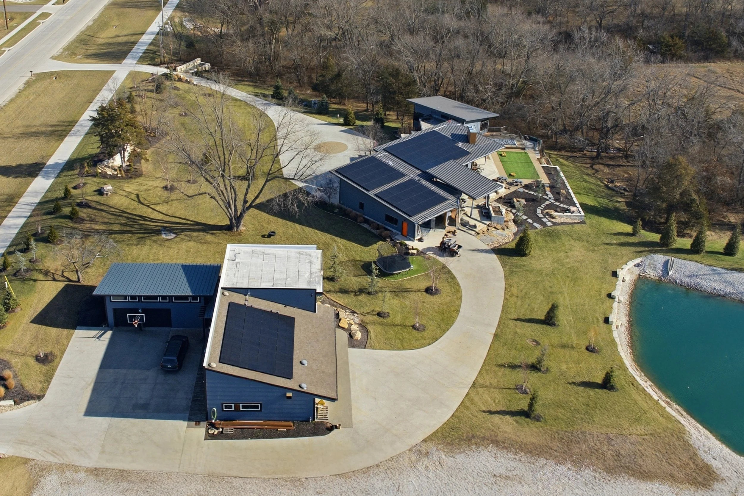 Aerial view of a modern house in Kansas with solar panels on the roof, surrounded by a landscaped yard, trees, and a nearby pond. Energy independence. 