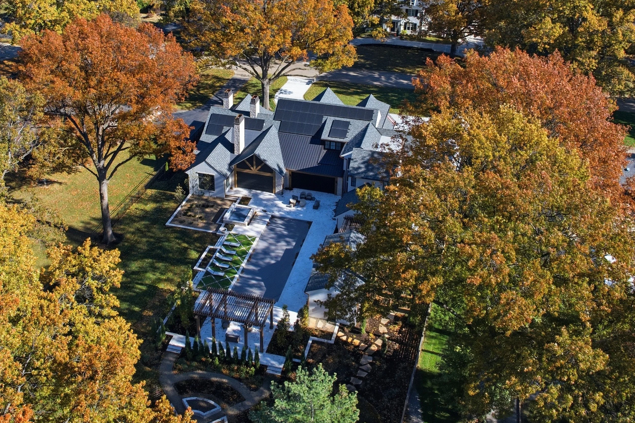 Aerial view of a house surrounded by fall trees with orange and yellow leaves, featuring a tiled roof with solar panels, paved walkways, and landscaped gardens.