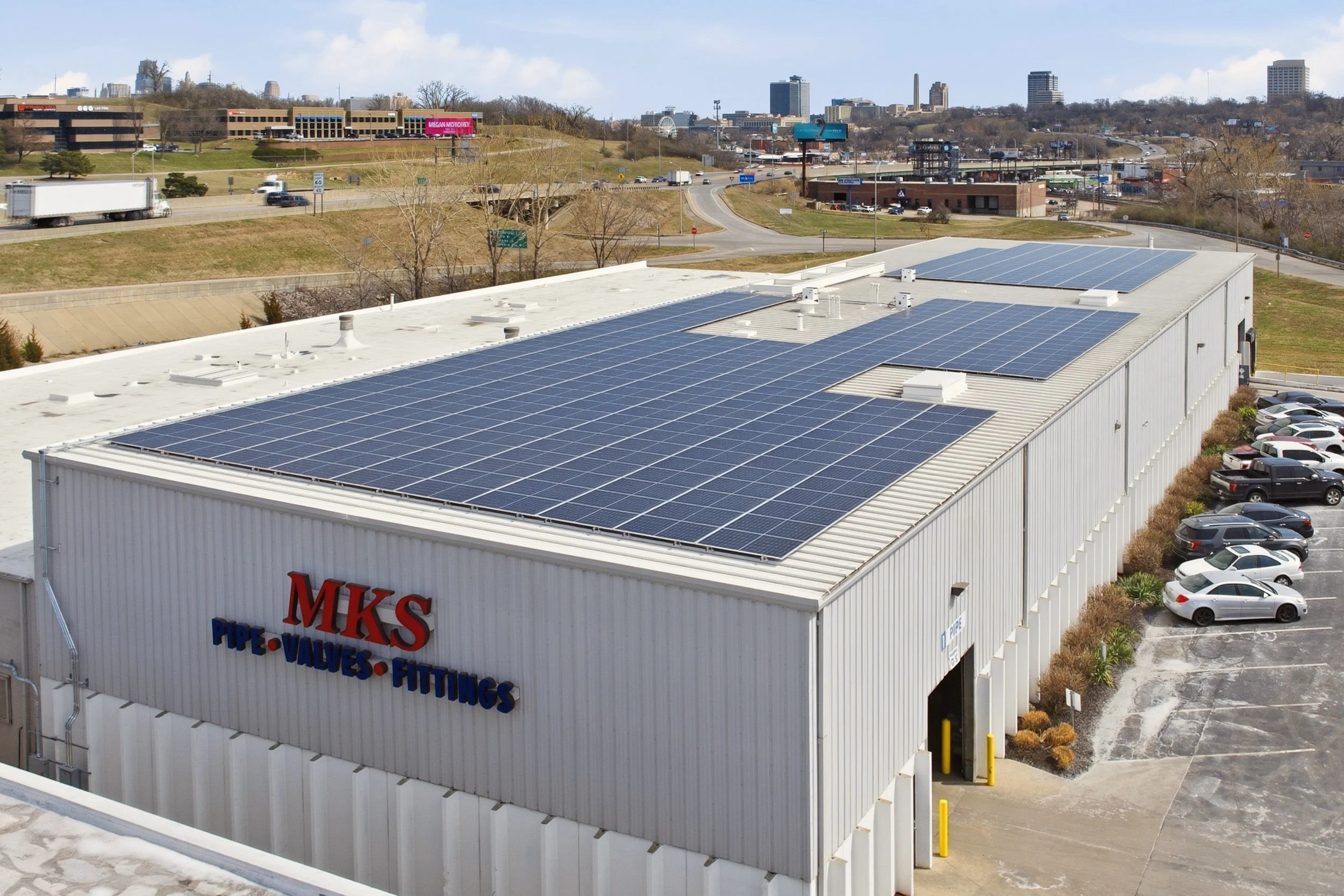 A commercial building in Kansas City, Kansas with solar panels on its white roof, part of a parking lot with cars, and a city skyline in the background.