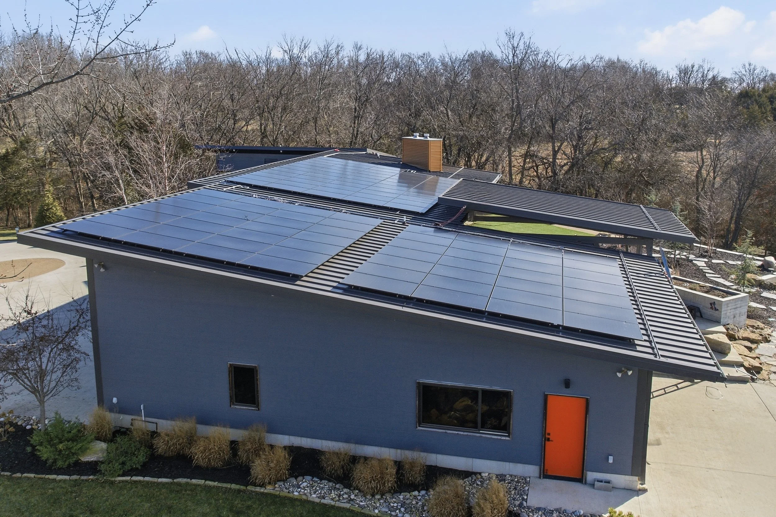 Aerial view of a modern house in Overland Park, Kansas with 60kW of solar panels on the roof, surrounded by leafless trees and landscaping. Sustainable, eco-friendly living.
