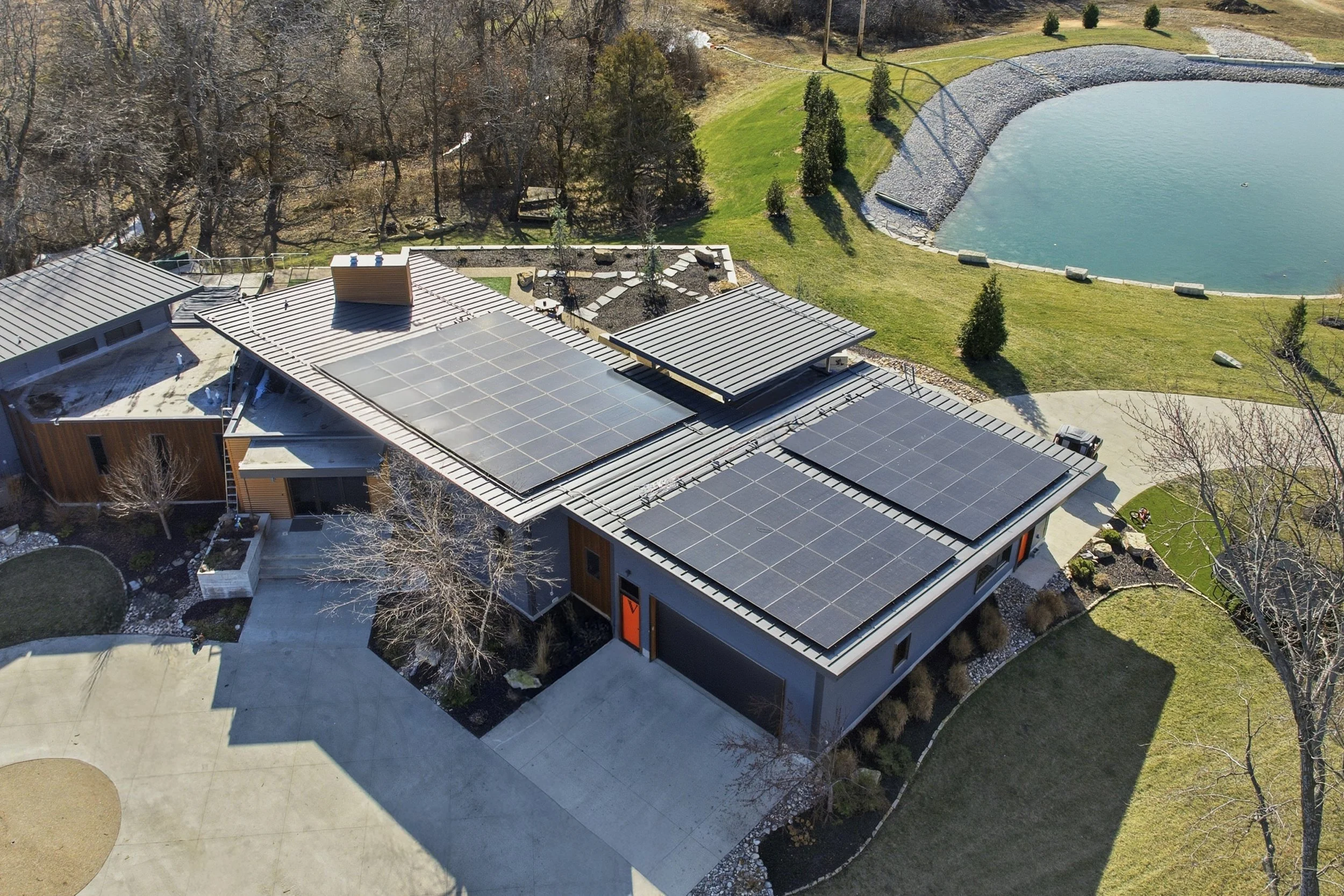 Aerial view of a modern house in Johnson County, Kansas with 60kW of solar panels on the roof, a backyard with a pond, trees, and landscaping in a suburban neighborhood. The homeowner is living sustainably with eco-friendly design. 