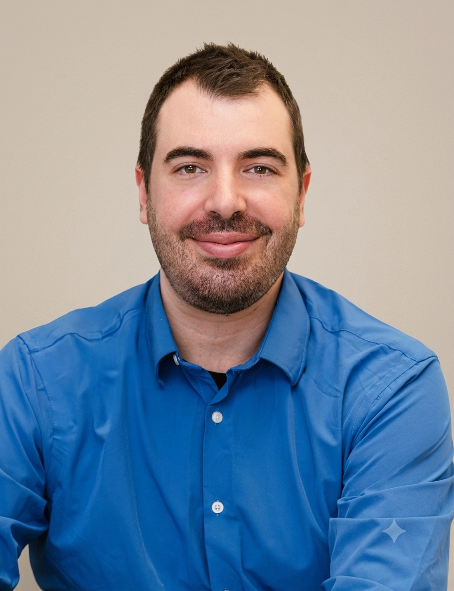 Close-up of a man with brown hair and a beard, smiling while wearing a blue collared shirt, sitting indoors against a neutral background.