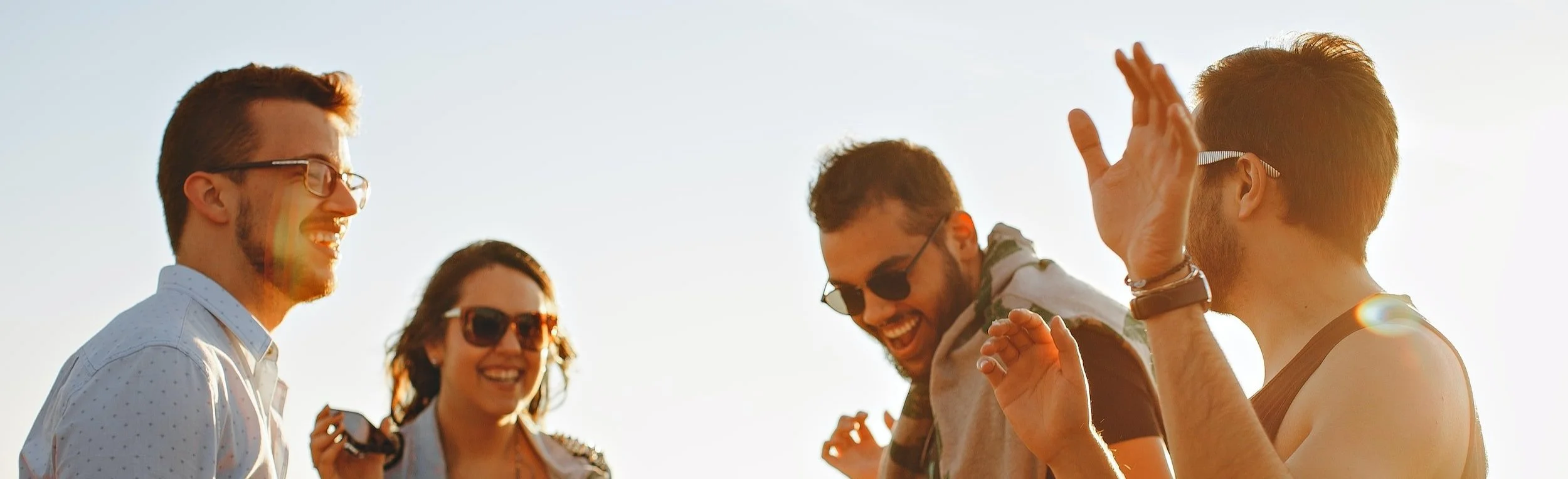 Group of four friends laughing outdoors in bright sunlight, wearing casual clothes and sunglasses.