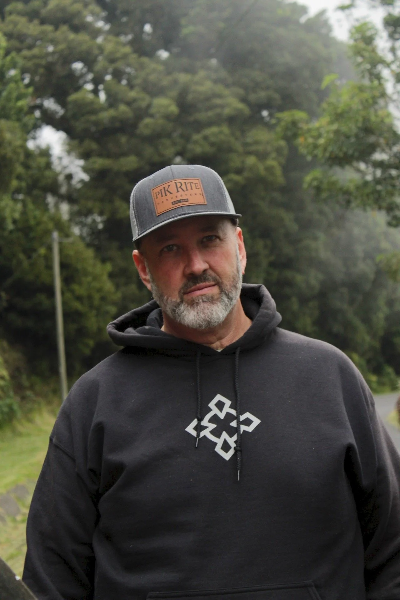 Smiling bald man with beard in plaid shirt outdoors with green trees in background.