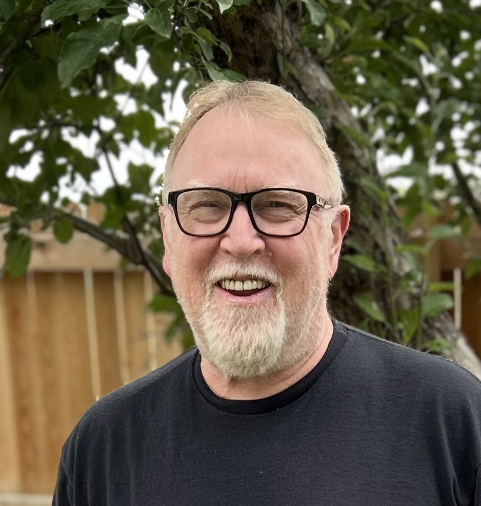 A man wearing glasses and a black shirt stands outdoors with green hills and a blue sky in the background.