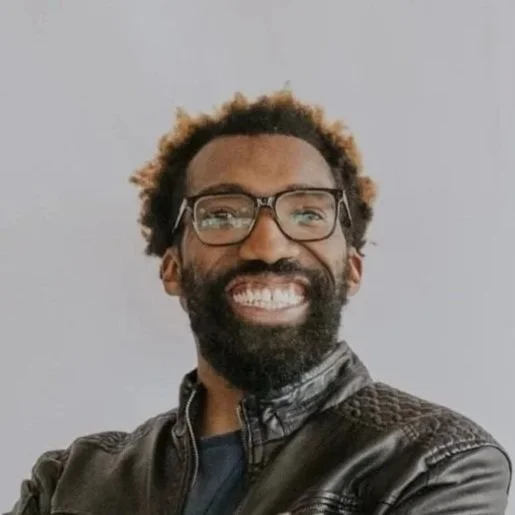Smiling man with glasses, beard, and curly hair, wearing a black leather jacket.