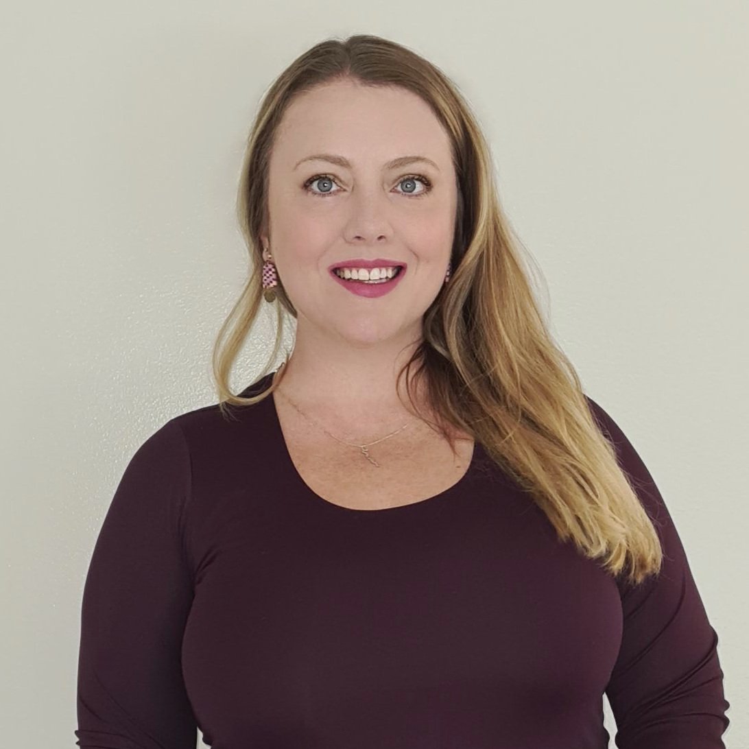 A woman with long, wavy strawberry blonde hair, wearing a dark long-sleeve top, pink patterned earrings, a necklace, and smiling, standing against a plain light-colored wall.