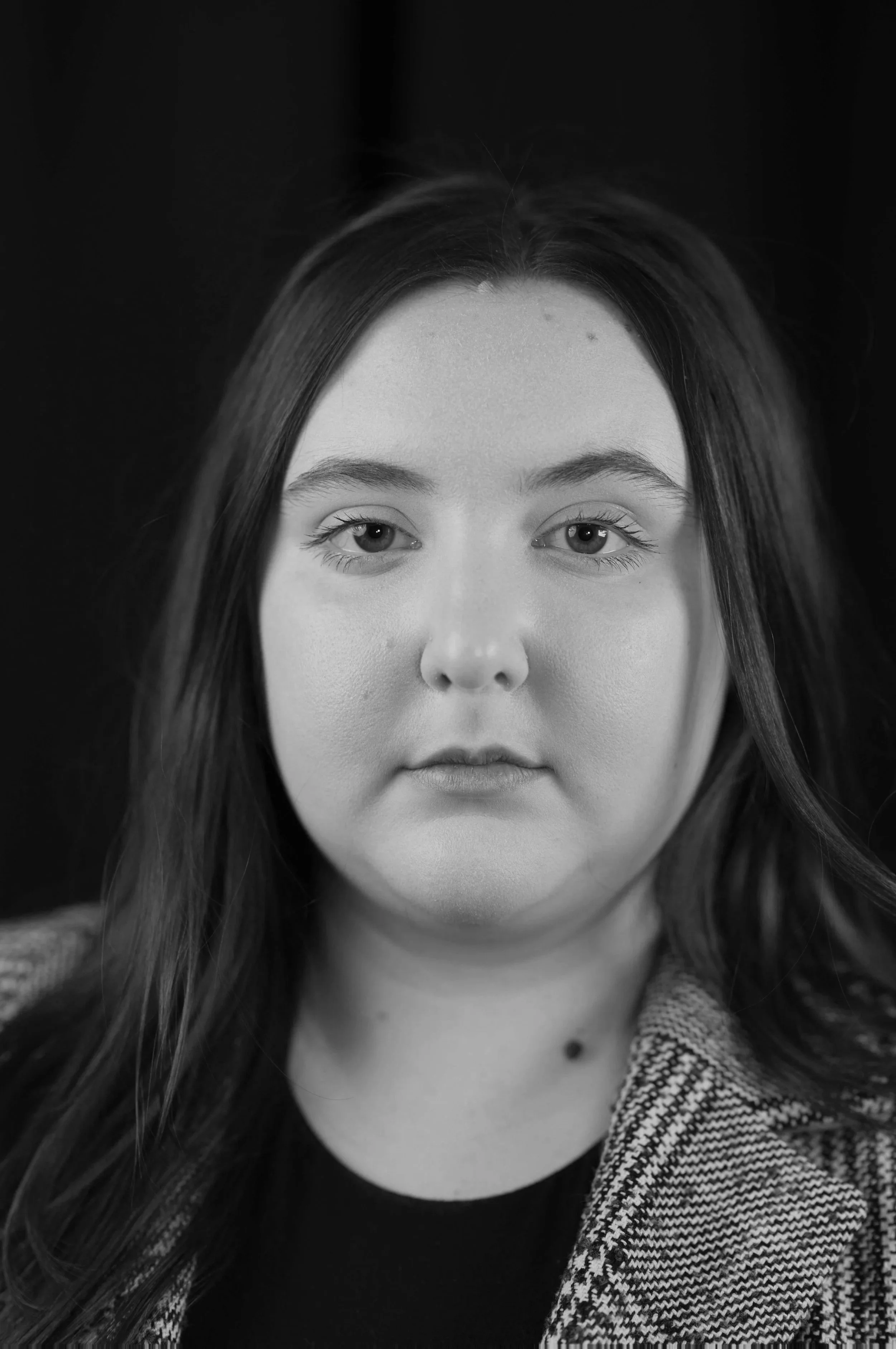 A black and white photo of a woman with long hair, looking directly at the camera, with a dark background.