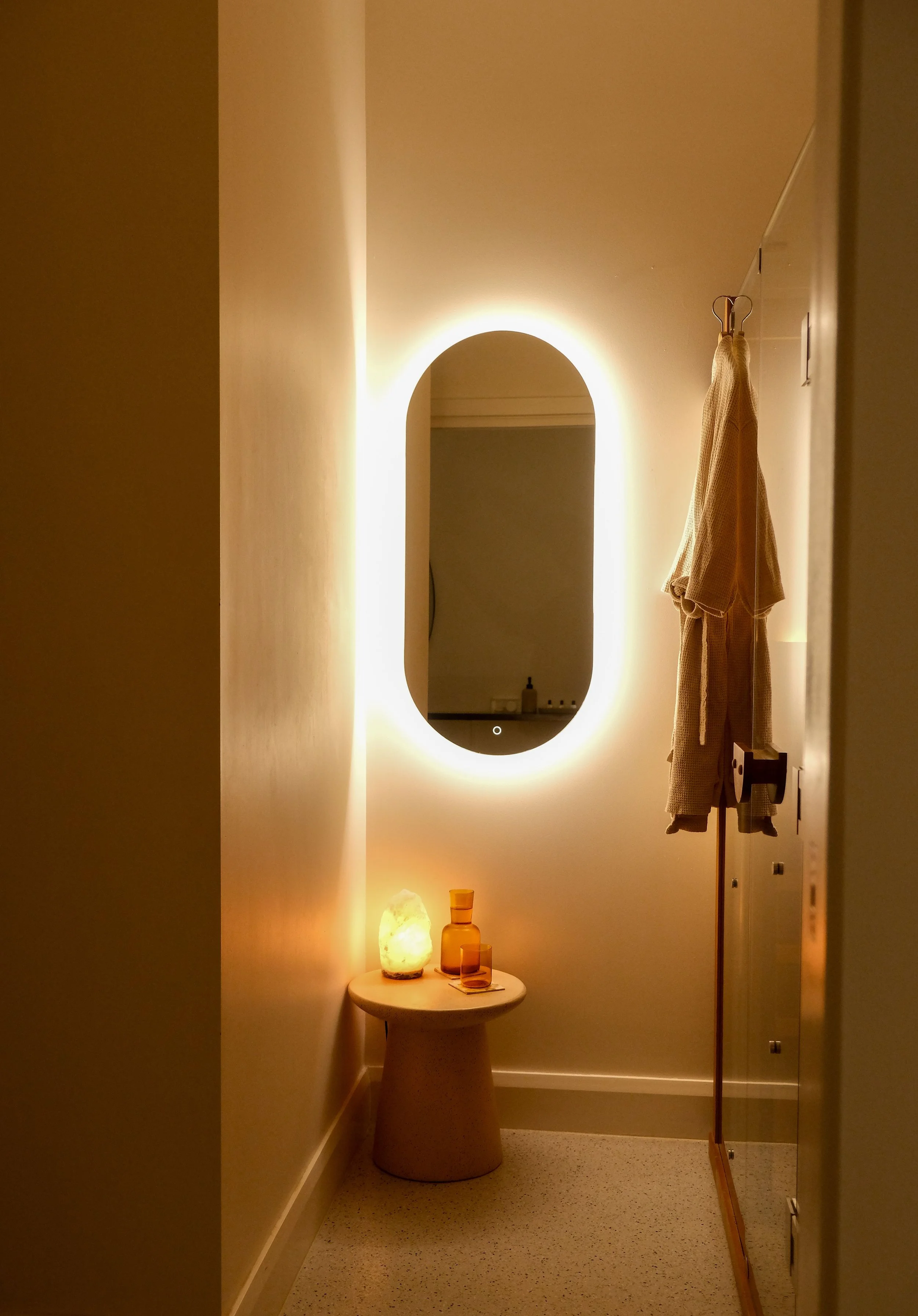 Warmly lit change room with backlit oval mirror and robe at Saltt Burleigh Heads