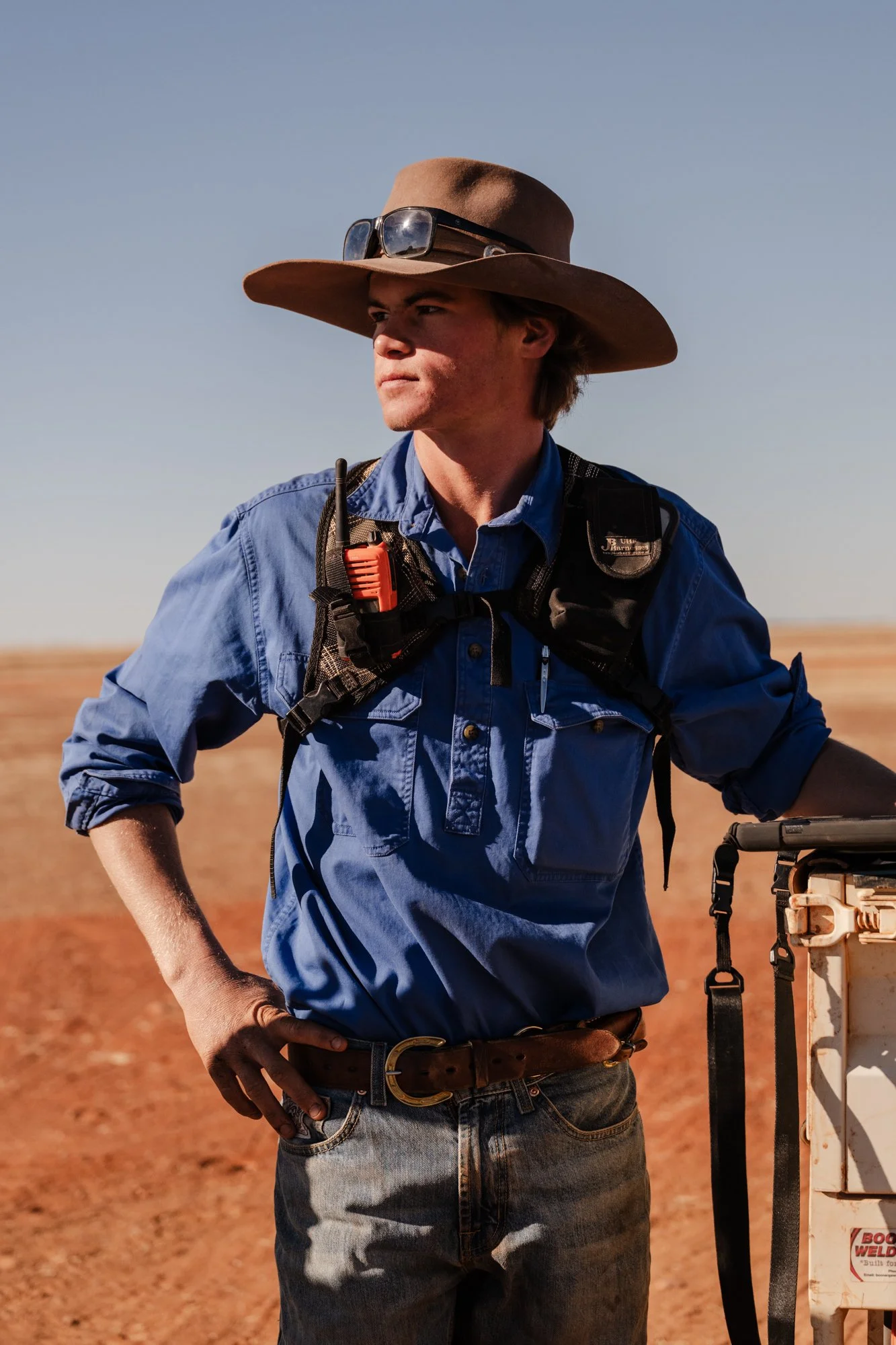 A young man wearing a wide-brimmed hat, sunglasses, a blue button-up shirt, jeans, and a harness with a communication device, standing outdoors with a clear sky and earthy terrain in the background.