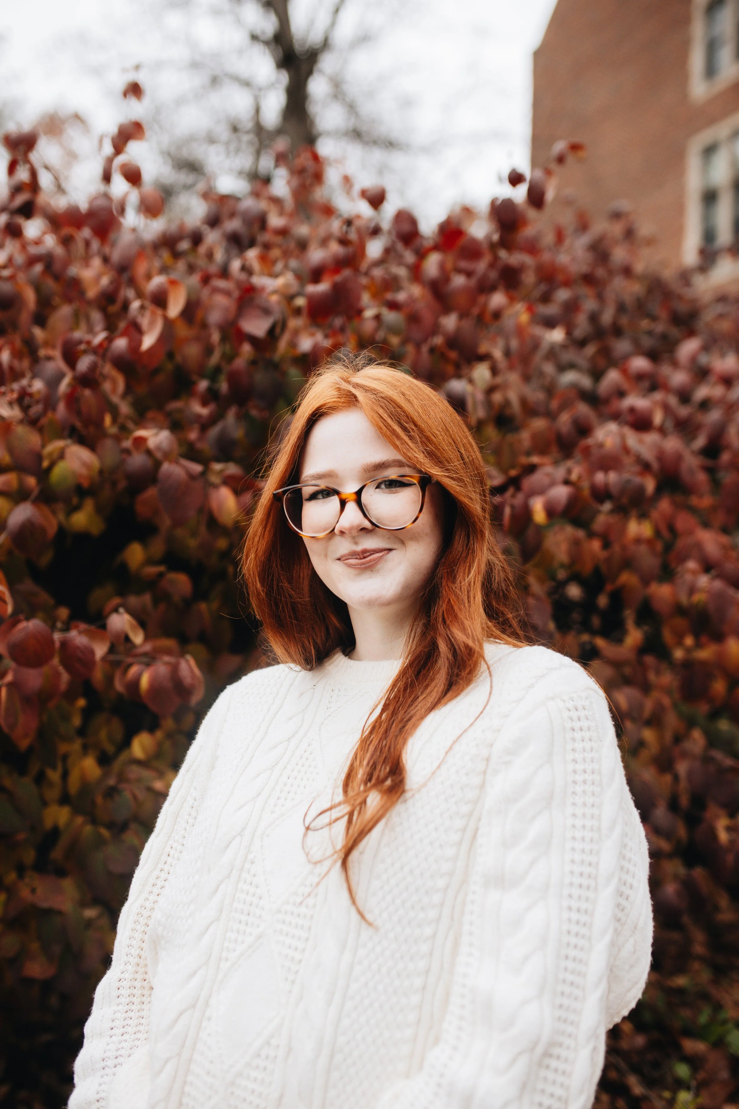 A young woman with long red hair, glasses, and freckles, smiling in a field of yellow and orange flowers with green trees in the background.