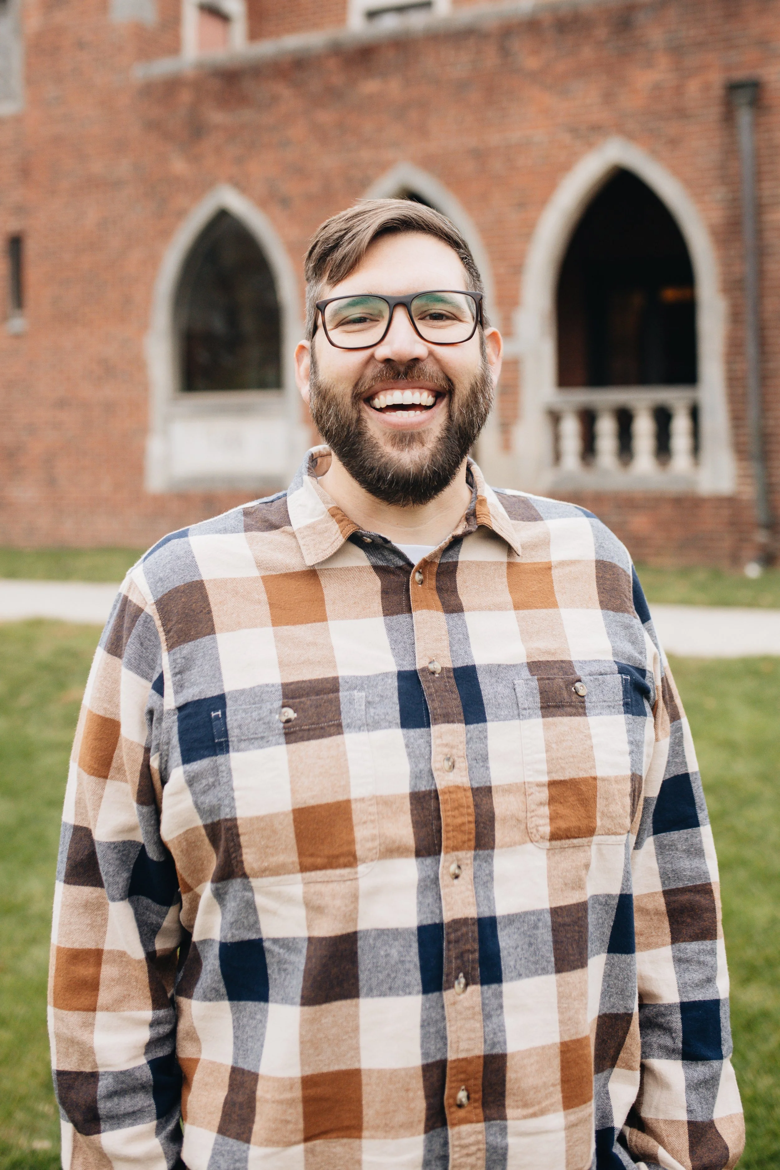 A man with glasses, a beard, and a checkered shirt smiling outdoors with trees in the background.