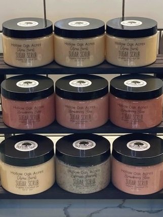 A collection of goat milk sugar scrubs in clear jars with black lids displayed on a tiered black shelf.