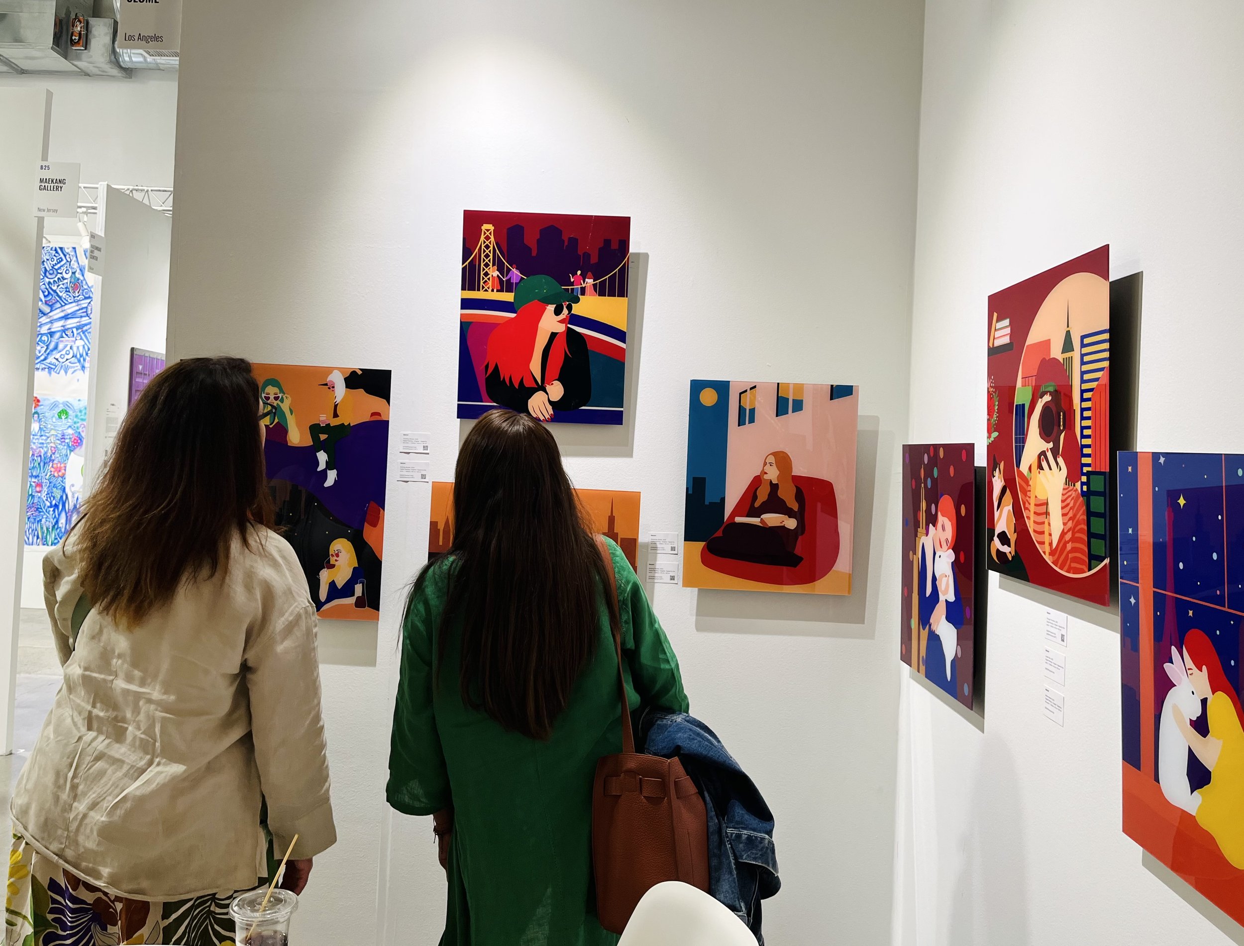 Two women viewing colorful digital art pieces in a gallery, featuring vibrant illustrations of people in various settings.