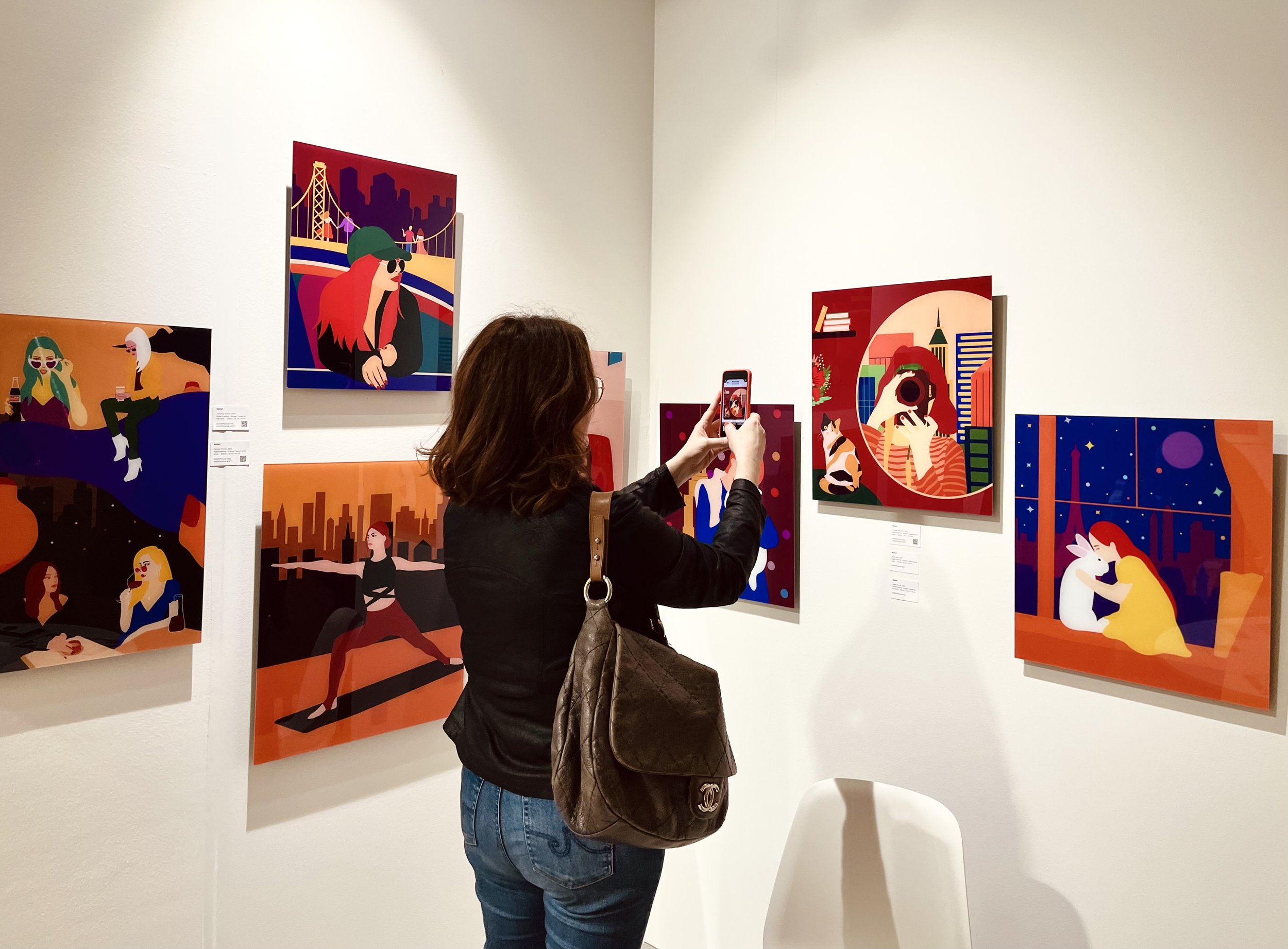 Art gallery exhibition with colorful, modern illustrations on display. A woman with brown hair is taking a photo of the artwork using her smartphone. She is wearing a black jacket and carrying a gray shoulder bag. The artworks depict various scenes w