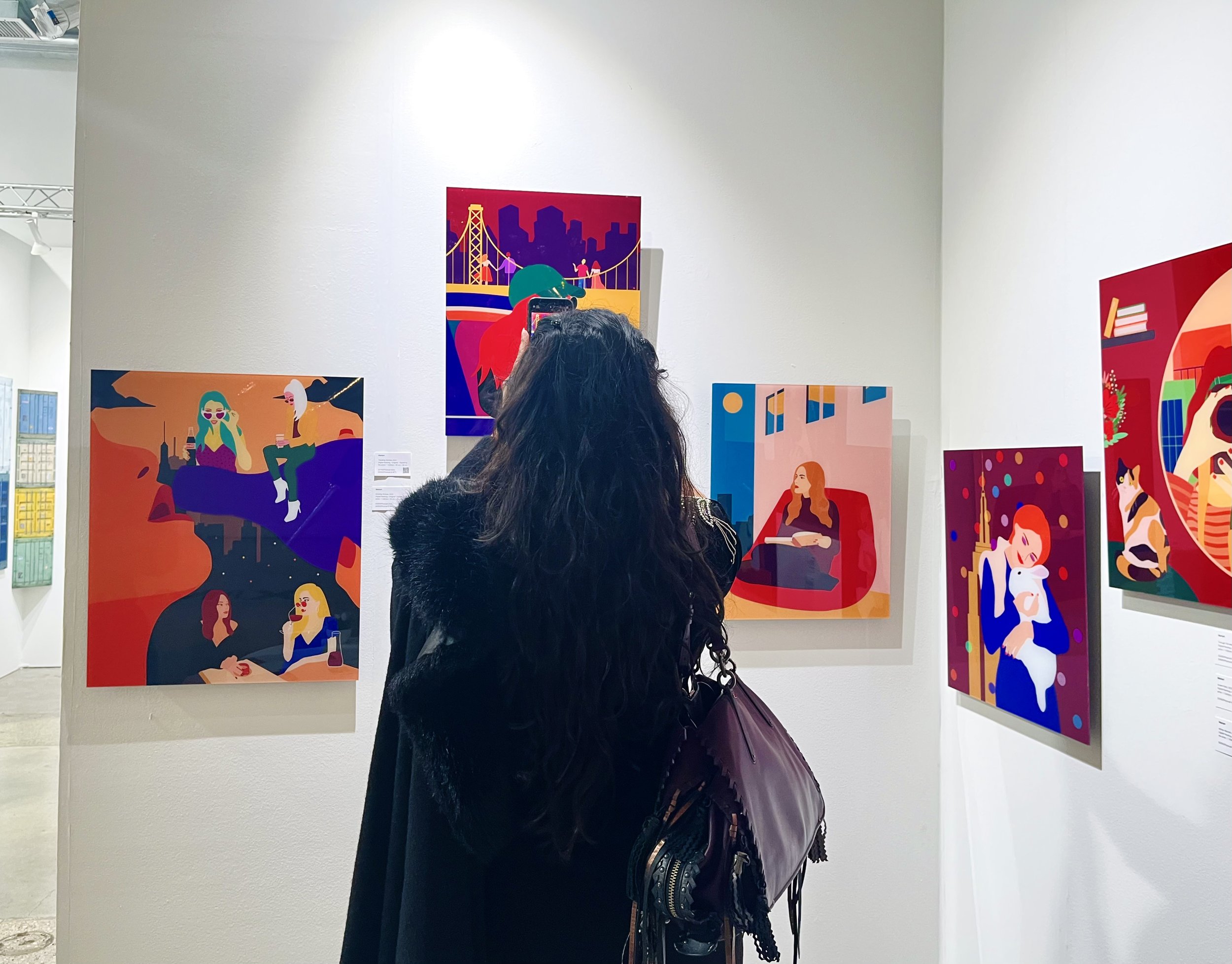 A person with long dark hair stands in front of a wall displaying colorful, modern art paintings in a gallery. The artworks feature abstract and stylized human figures in vibrant colors and depict various scenes, such as a cityscape with a bridge, pe