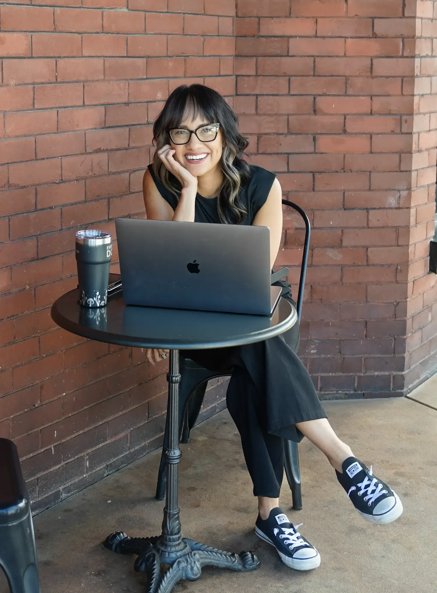 Image of woman with dark hair and glasses sitting at a cafe table with a laptop and coffee