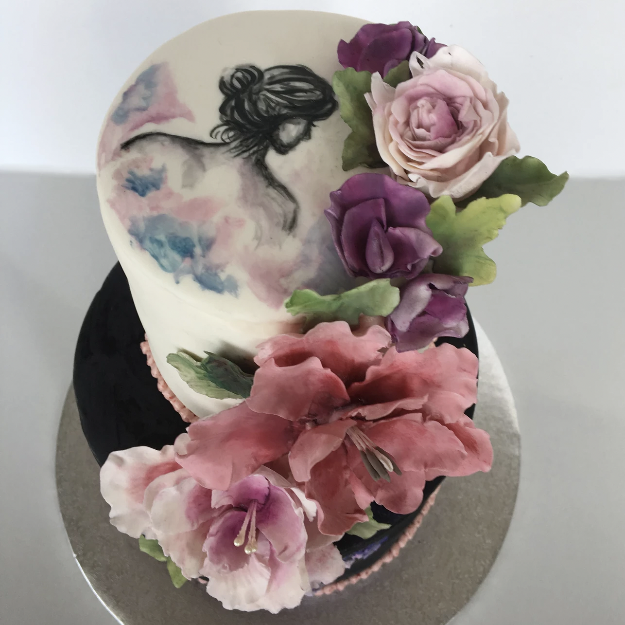 Decorated cake with painted silhouette of a woman and colorful sugar flowers, including roses and other blooms, on a white icing base.