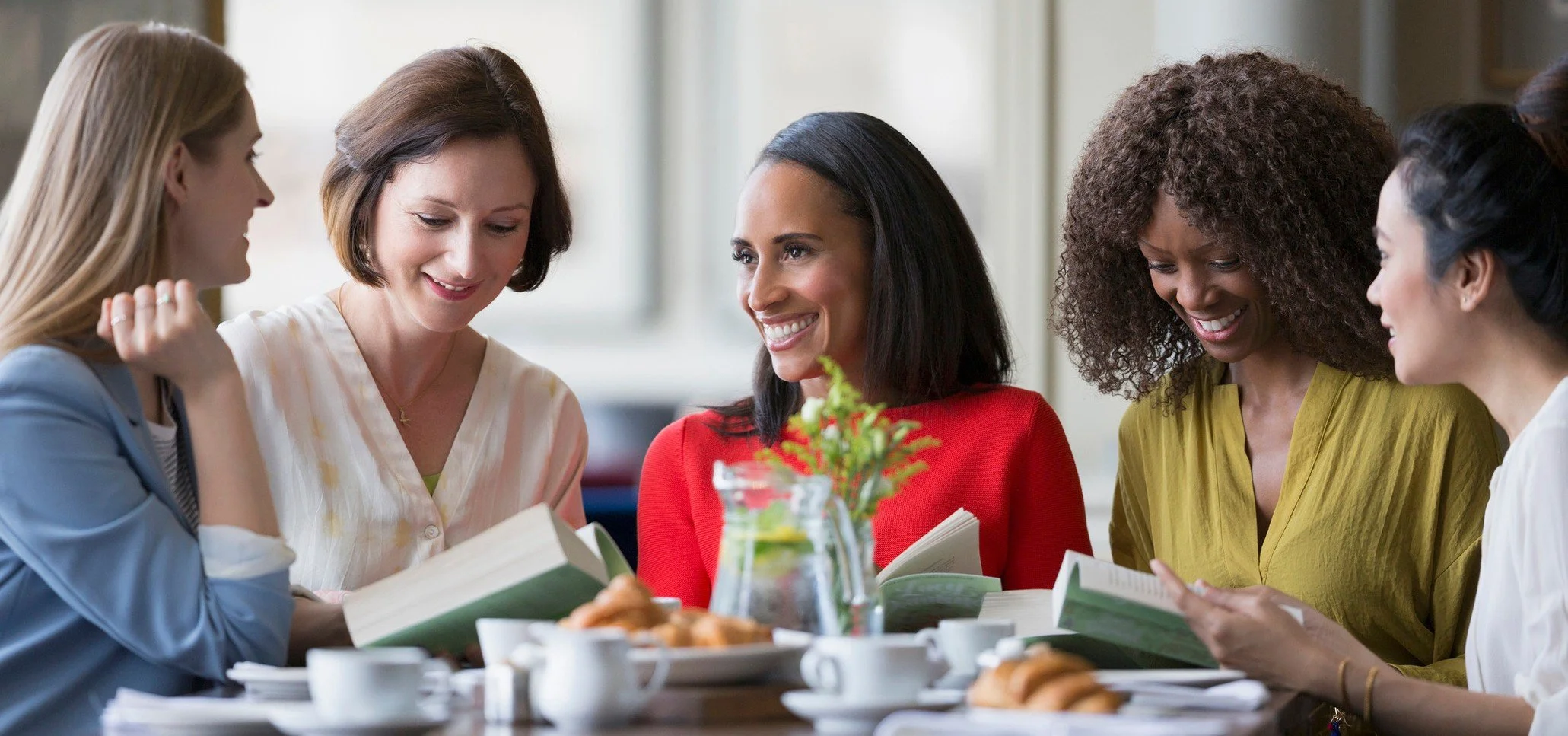 A group of five women gathered around a table over brunch with books in hand.