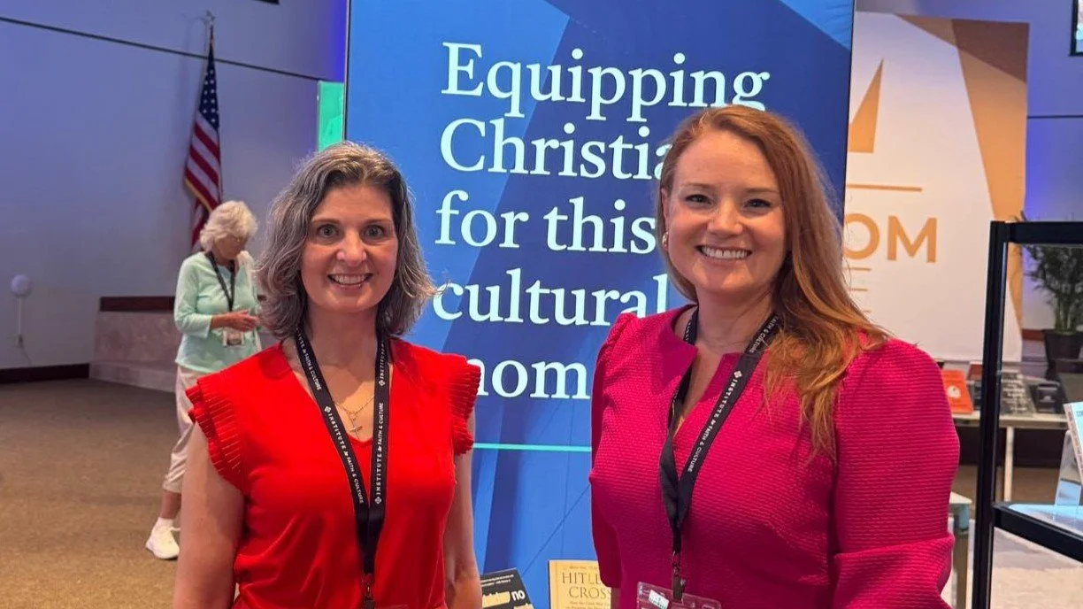 A women duo wearing red hues and standing in front of a blue retractable banner.