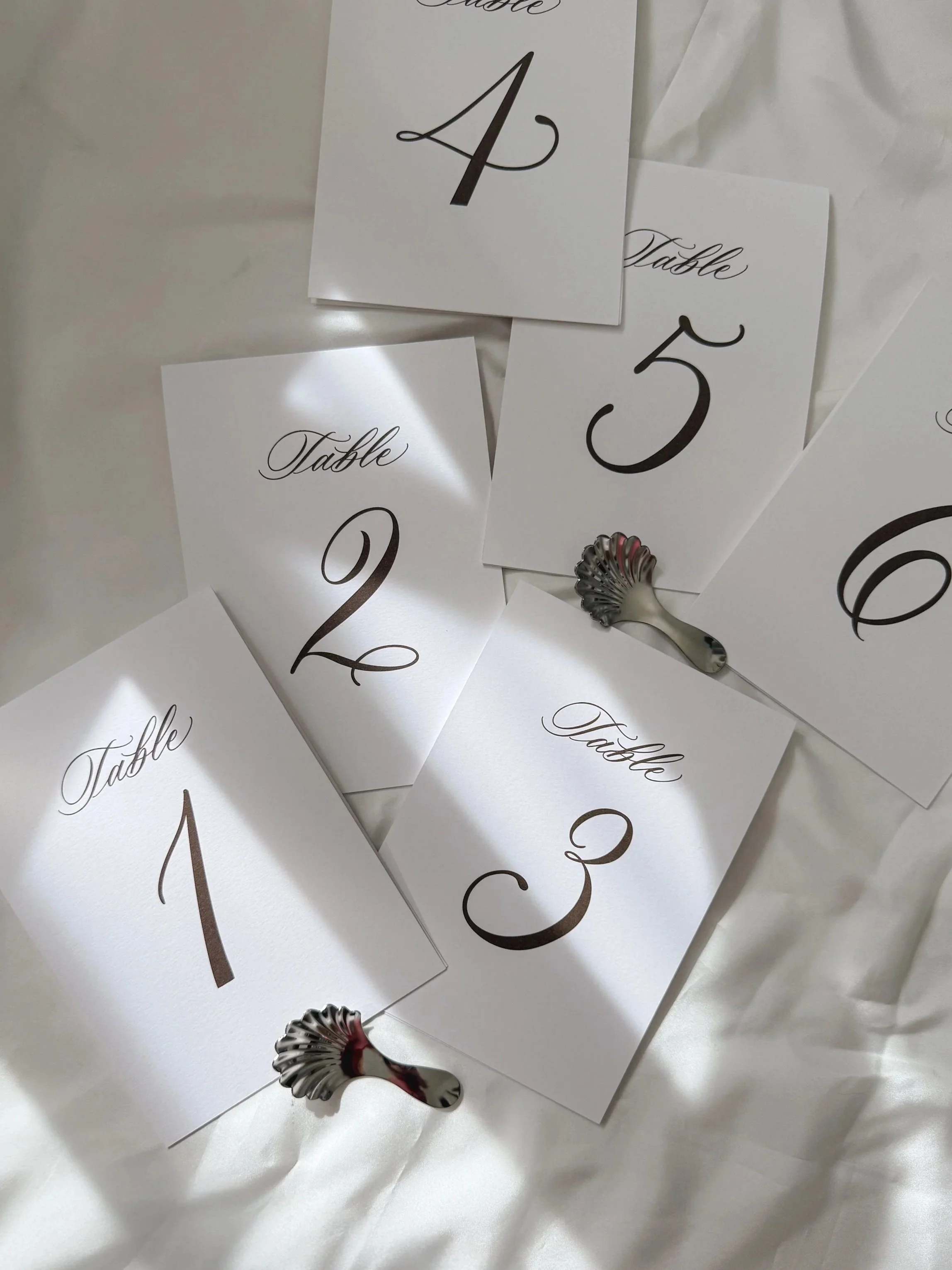 White cards with black cursive text displaying table numbers 1, 2, 3, 4, 5, and 6, scattered on a white fabric surface, with two metal table number holders.