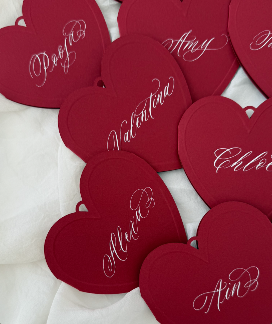 Red heart-shaped cards with white cursive names written on them, scattered on a white surface.