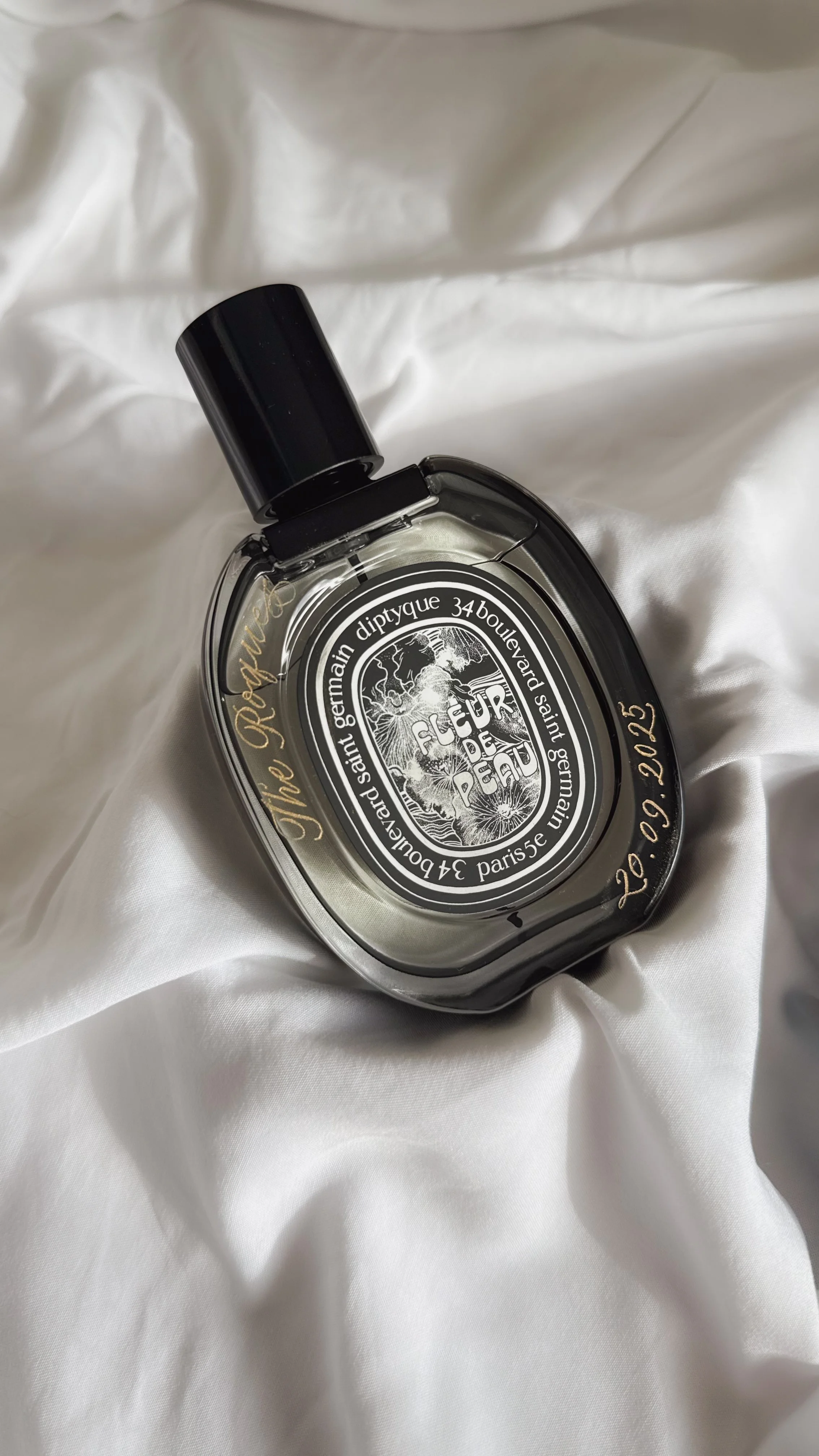 A transparent perfume bottle with black and gold accents, resting on white fabric. The label reads 'Fleur De Peau,' with handwritten gold text indicating 'V. No. 28.09.2025.'