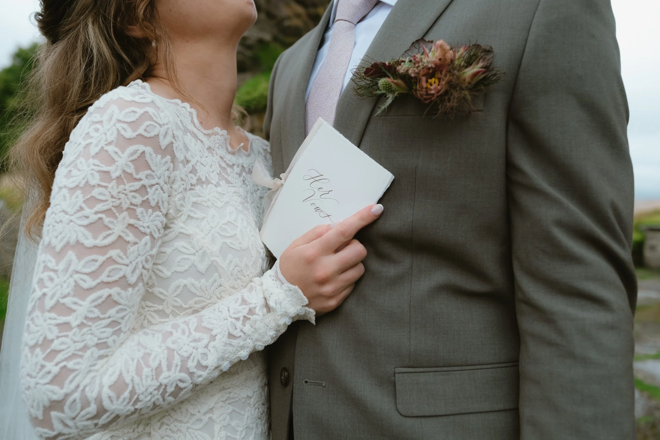 Close-up of a bride and groom during their wedding ceremony. The bride is holding a small white book that says 'Our Vows' and is wearing a white lace wedding dress. The groom is dressed in a light gray suit with a floral boutonniere on his lapel.