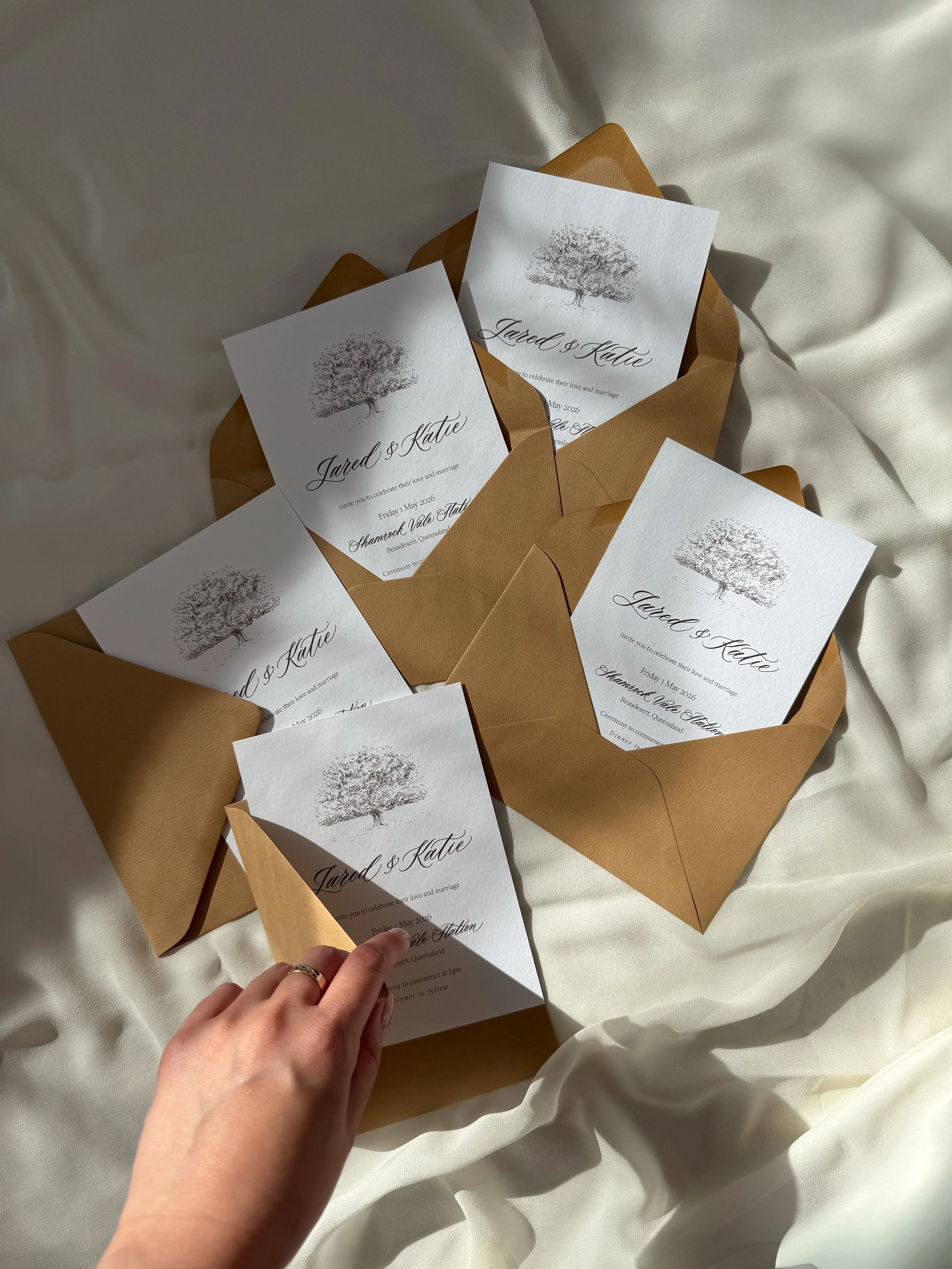Invitation cards for a wedding with a tree graphic, placed in brown envelopes on a cream-colored fabric background.