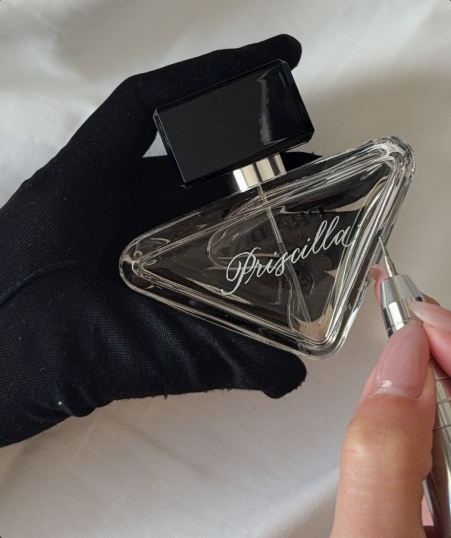 A hand wearing a black glove holds a Prada perfume bottle and is using a small tool to remove or adjust the label or cap.
