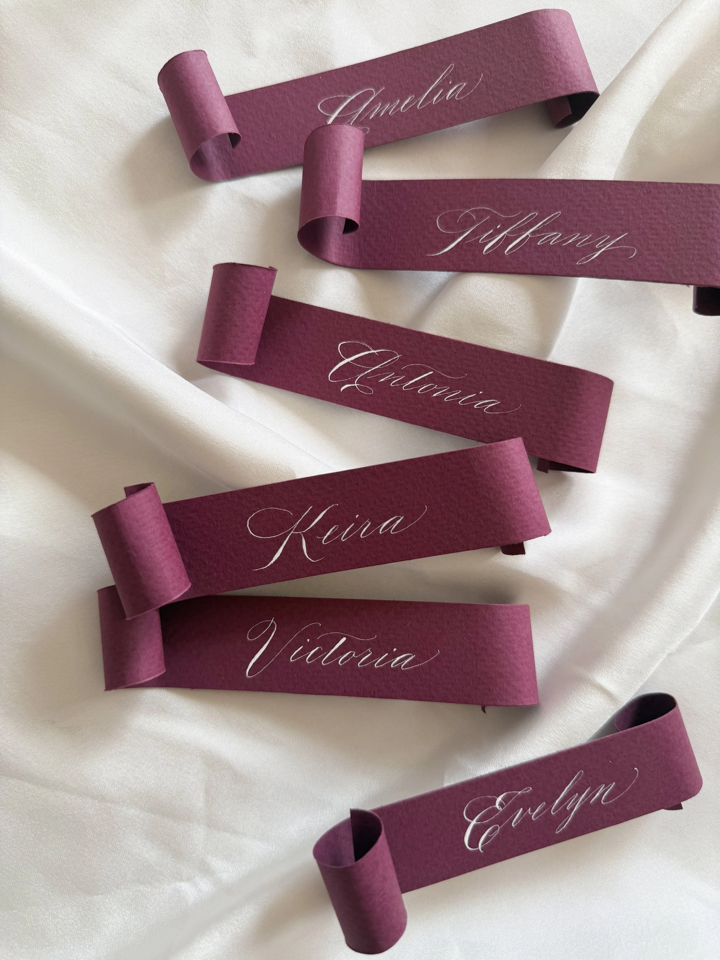 Multiple purple ribbon name tags with white cursive names, including Amelia, Tiffany, Augusta, Keira, Victoria, and Evelyn, arranged on a white fabric background.